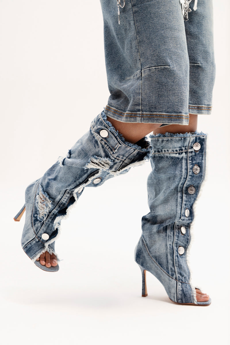 A close-up shows a person wearing the AZALEA WANG KAZUKI DENIM OPEN TOE BOOT, knee-high with heels, button details, and frayed edges. Paired with mid-length denim shorts, these chic boots feature a Comfort Cushion insole for style and comfort against a light-colored background.