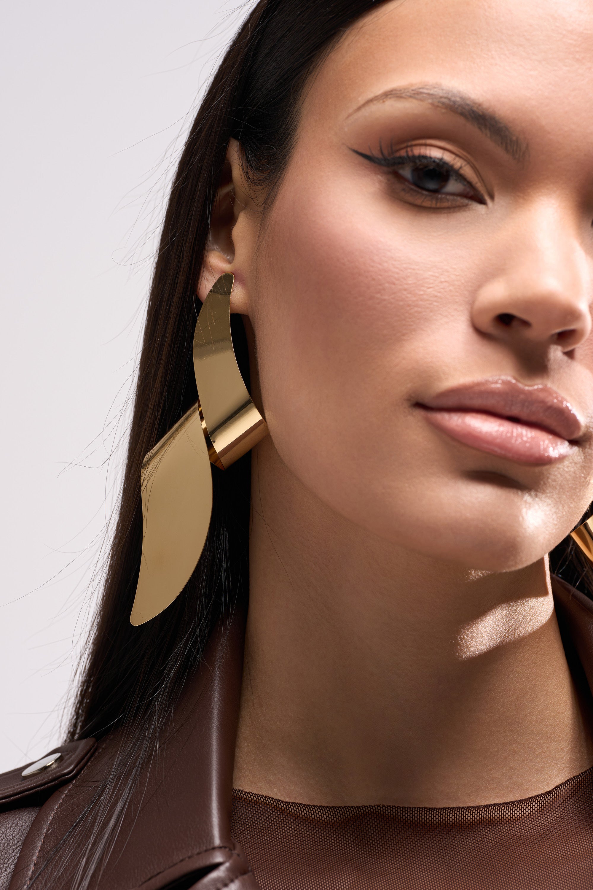 A woman with straight dark hair wears the TWISTED UP EARRING—a large, gold-tone, sculptural accessory—paired with a brown leather jacket. Her flawless makeup features winged eyeliner and nude lipstick, highlighting her earrings.