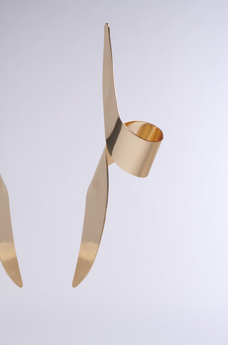 A close-up reveals a gold-colored foldable wall hook on a white backdrop, showcasing sleek, Twisted Up Earring-inspired design with sharp angles and a faux metallic gold finish for a modern aesthetic.