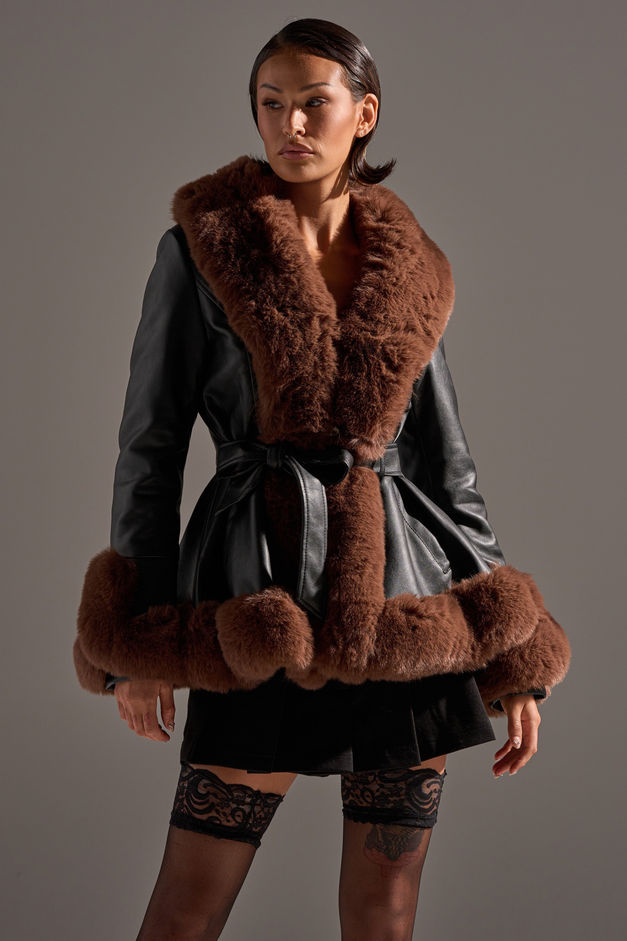 A woman wears the KENZY FUR TRIM PEPLUM MOTO IN BLACK BROWN, featuring bold brown faux fur on the collar, cuffs, and hem with a belted waist. She styles it with a short black skirt and lace stockings against a neutral background.