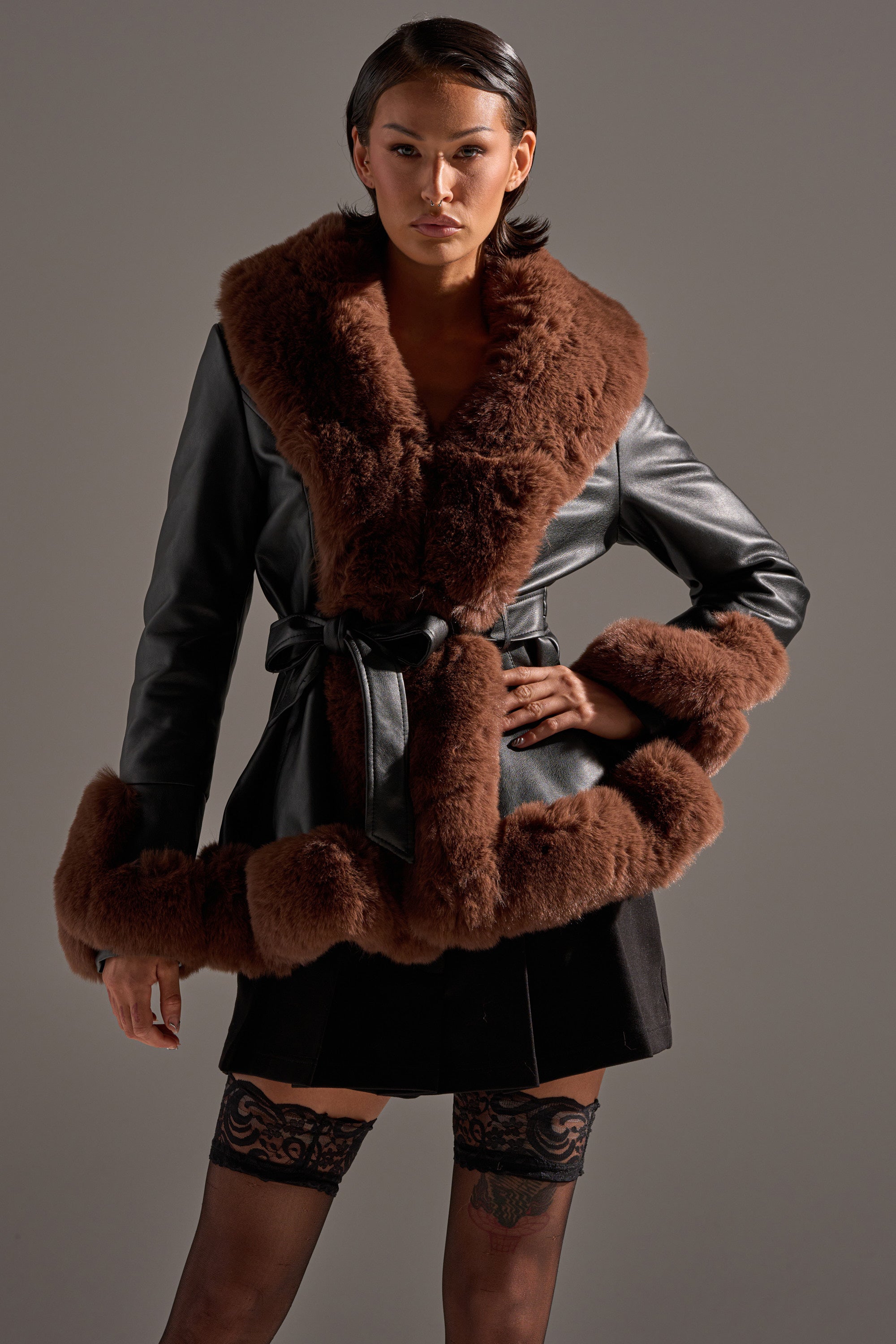 A woman exudes confidence in the KENZY FUR TRIM PEPLUM MOTO IN BLACK BROWN, featuring wide, plush brown faux fur accents on a black leather coat, styled with a short black skirt and black lace thigh-high stockings.