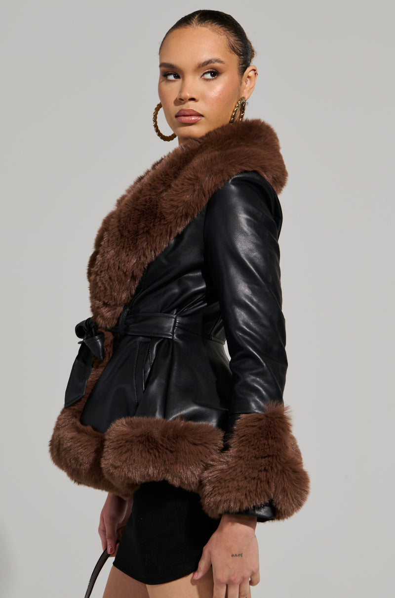 A person wearing hoop earrings sports the KENZY FUR TRIM PEPLUM MOTO IN BLACK BROWN, a black leather jacket with faux fur trim and a belted waist. The peplum moto-inspired design adds a chic touch against the plain, light gray background.