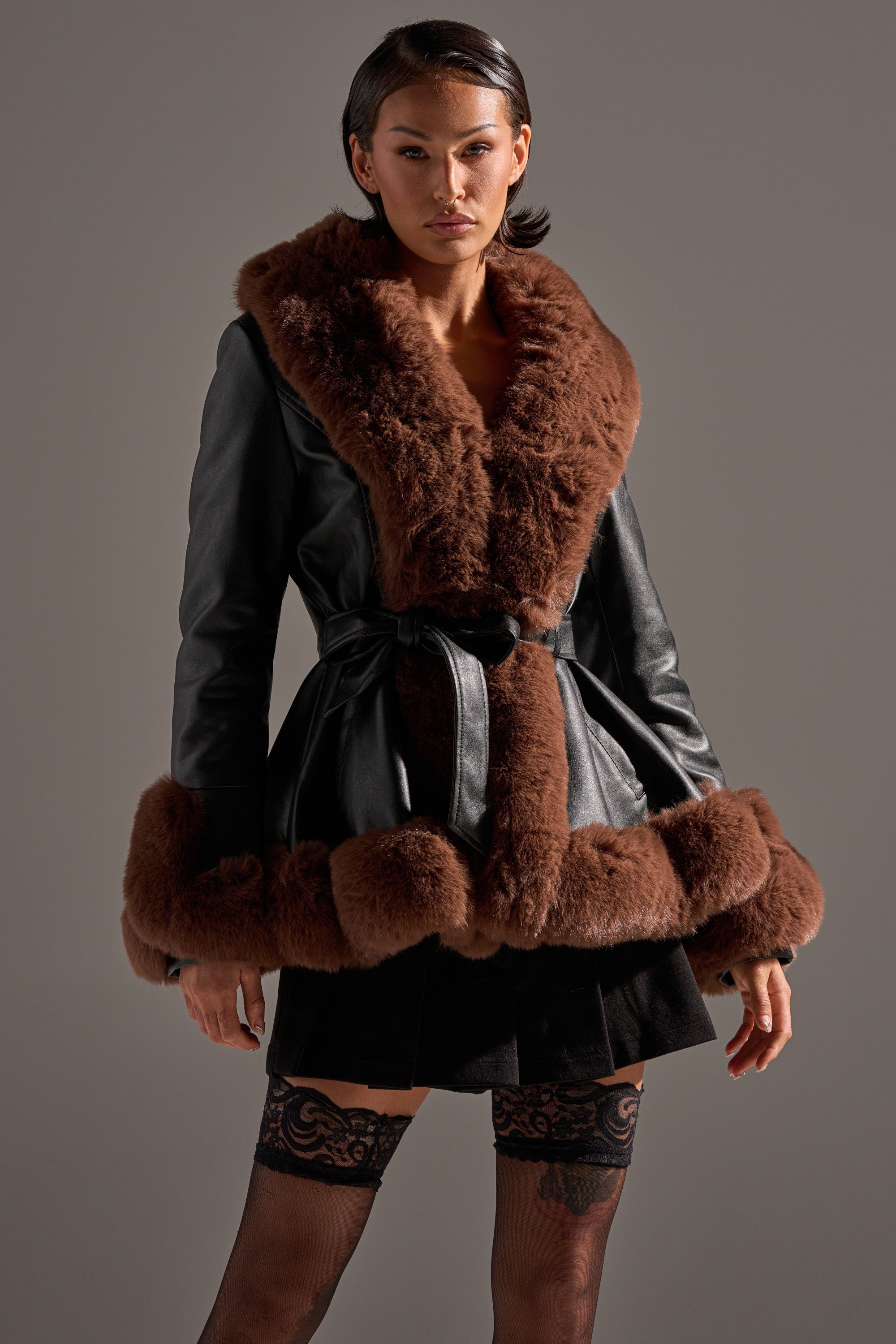 A woman wears the KENZY FUR TRIM PEPLUM MOTO IN BLACK BROWN, featuring thick brown faux fur on the collar, cuffs, and hem, paired with a black pleated skirt and black lace stockings, standing against a plain gray background.
