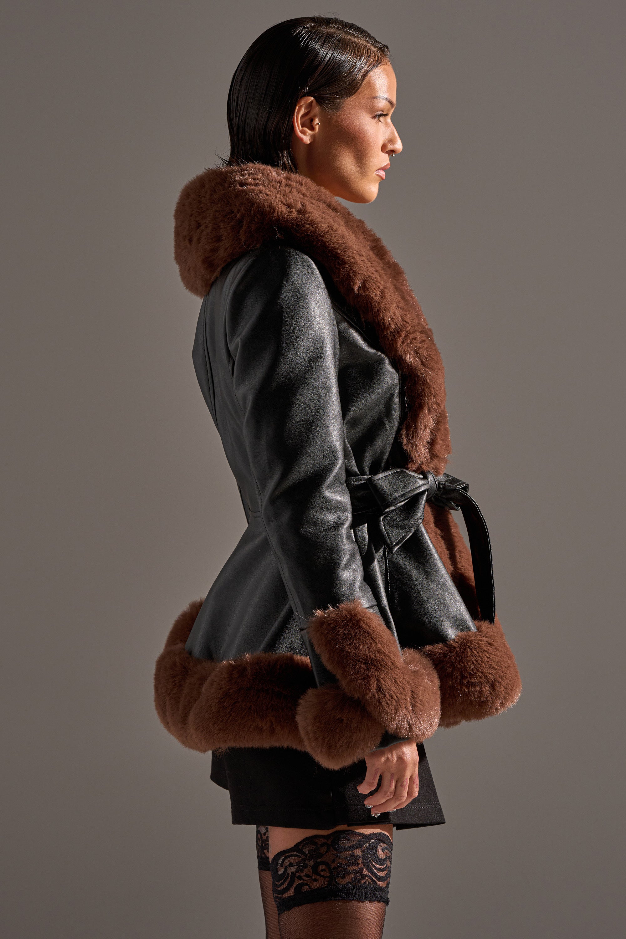 A woman in profile wears the KENZY FUR TRIM PEPLUM MOTO IN BLACK BROWN, featuring a belted silhouette with thick brown faux fur trim on the collar, cuffs, and hem, styled with a short black skirt and patterned lace stockings against a plain background.