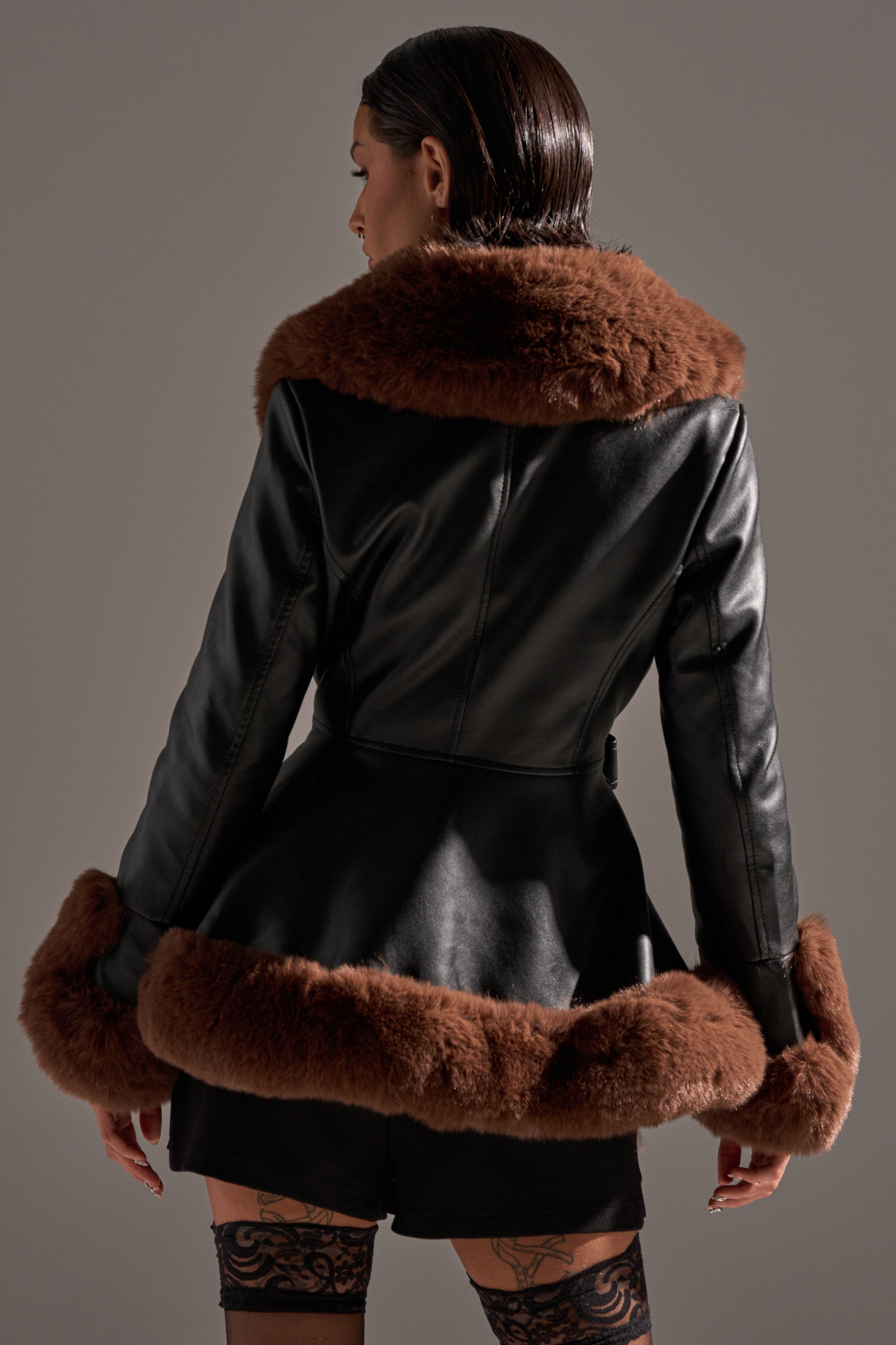 A woman faces away from the camera in the KENZY FUR TRIM PEPLUM MOTO IN BLACK BROWN, featuring brown faux fur trim at the collar, cuffs, and hem. Her slicked-back hair and lace stockings add an edgy touch beneath the jacket.