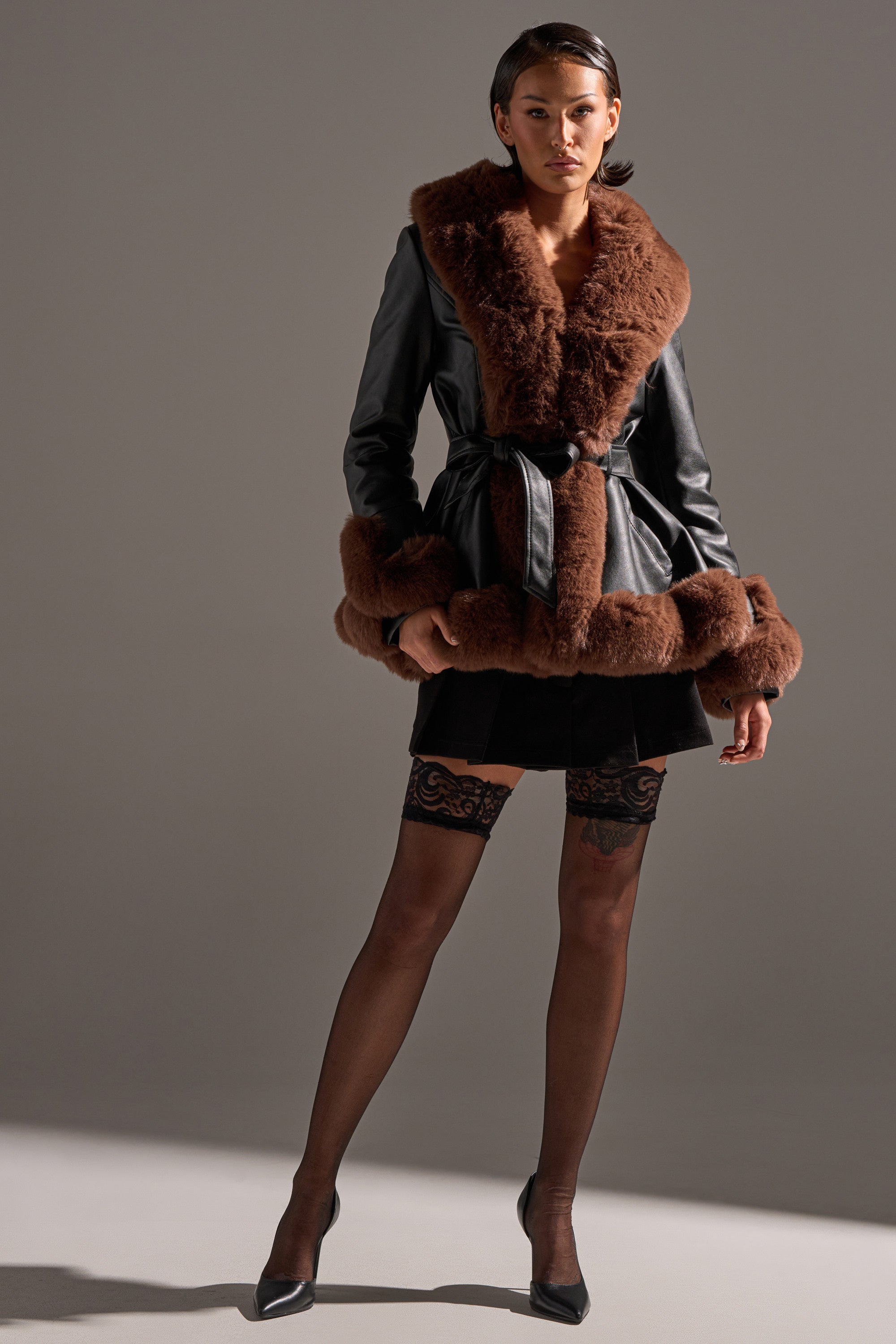 A woman stands confidently in the KENZY FUR TRIM PEPLUM MOTO IN BLACK BROWN, styled with a short black skirt, black lace-top stockings, and black heels against a plain gray background.