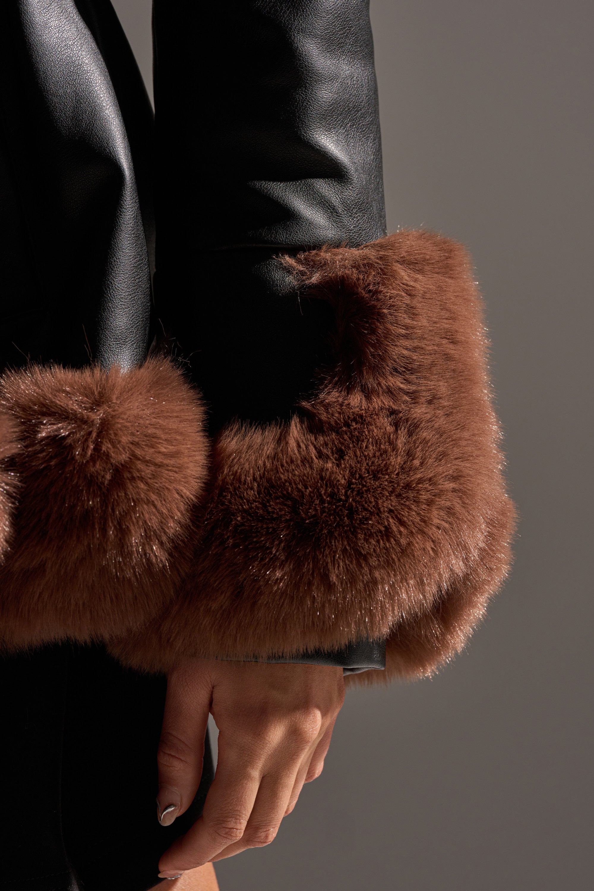 A close-up of a hand and arm wearing the KENZY FUR TRIM PEPLUM MOTO IN BLACK BROWN jacket with thick brown cuffs, set against a neutral background.