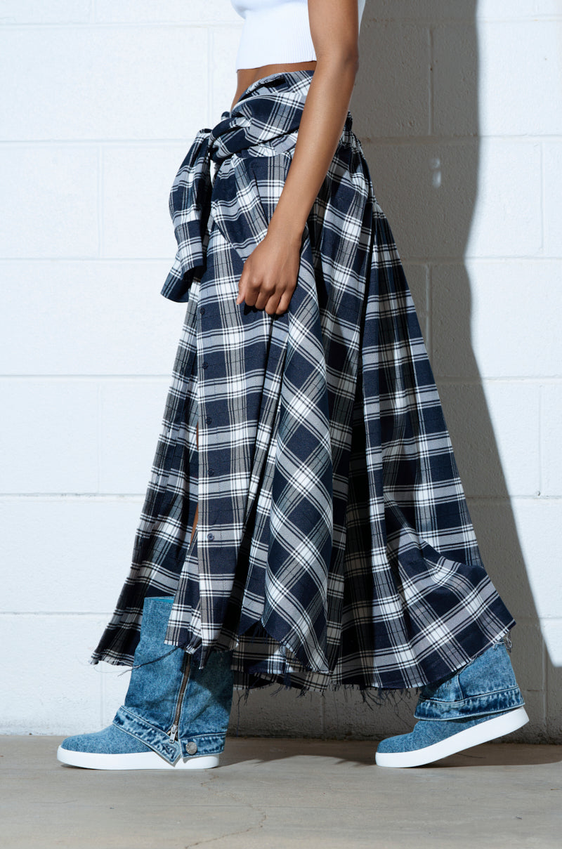 A person wearing a blue plaid skirt, a tied plaid shirt, and AZALEA WANG KIMIKA DENIM BOOTIE strides past a white brick wall, exuding a casual yet stylish vibe.
