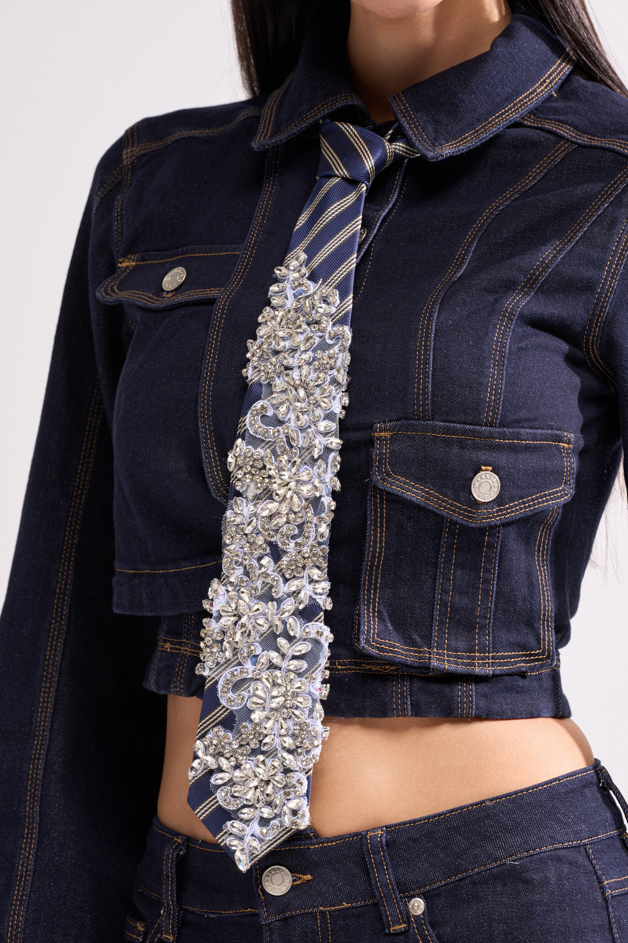 A woman sports a dark denim cropped jacket and jeans, complemented by NOT YOUR TYPICAL TIE—a blue striped rhinestone accessory that adds flair to her business look. Her face is partially out of the frame.