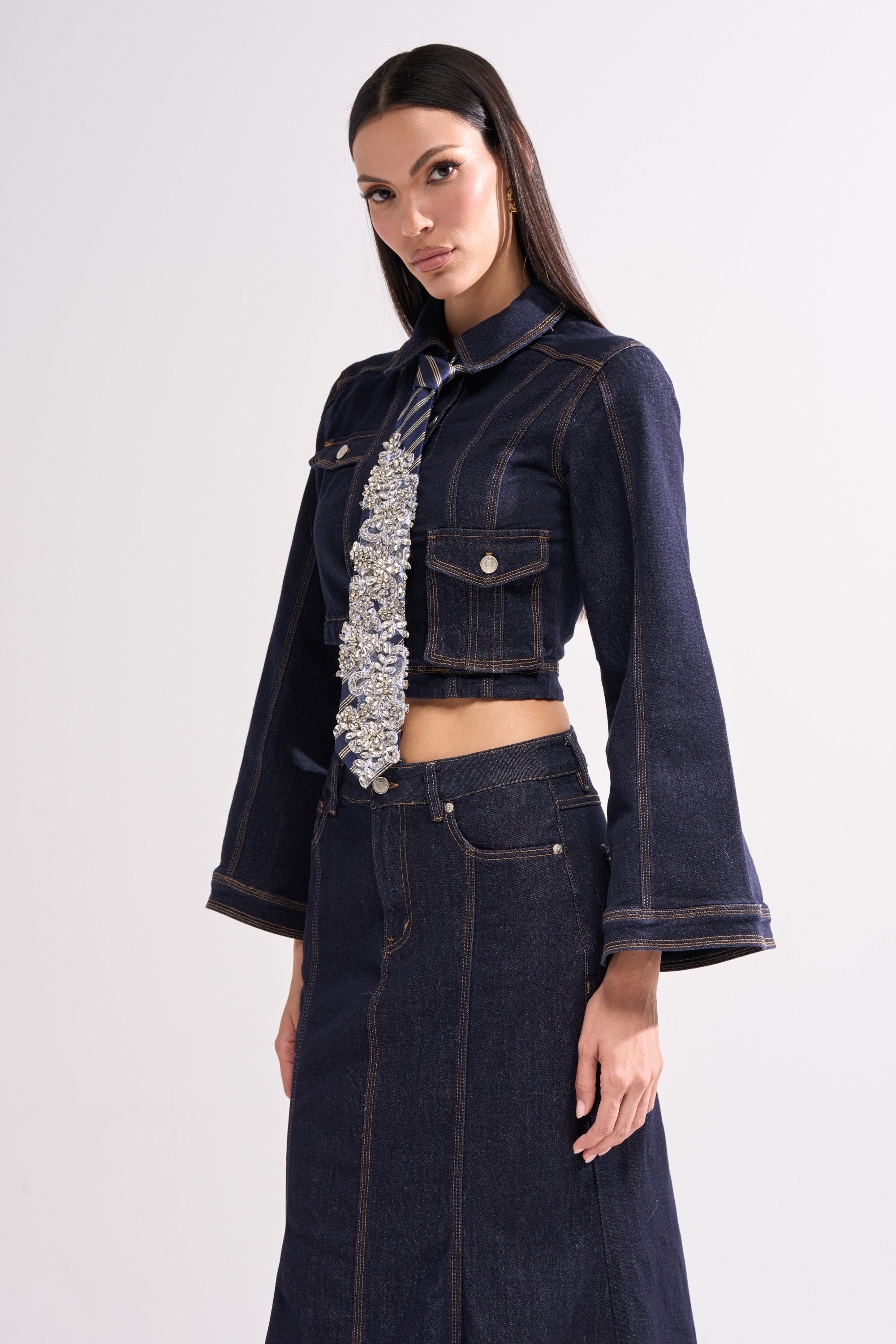 A woman wears a dark denim cropped jacket with flared sleeves, a matching high-waisted skirt, and the NOT YOUR TYPICAL TIE rhinestone tie, standing against a plain light background.