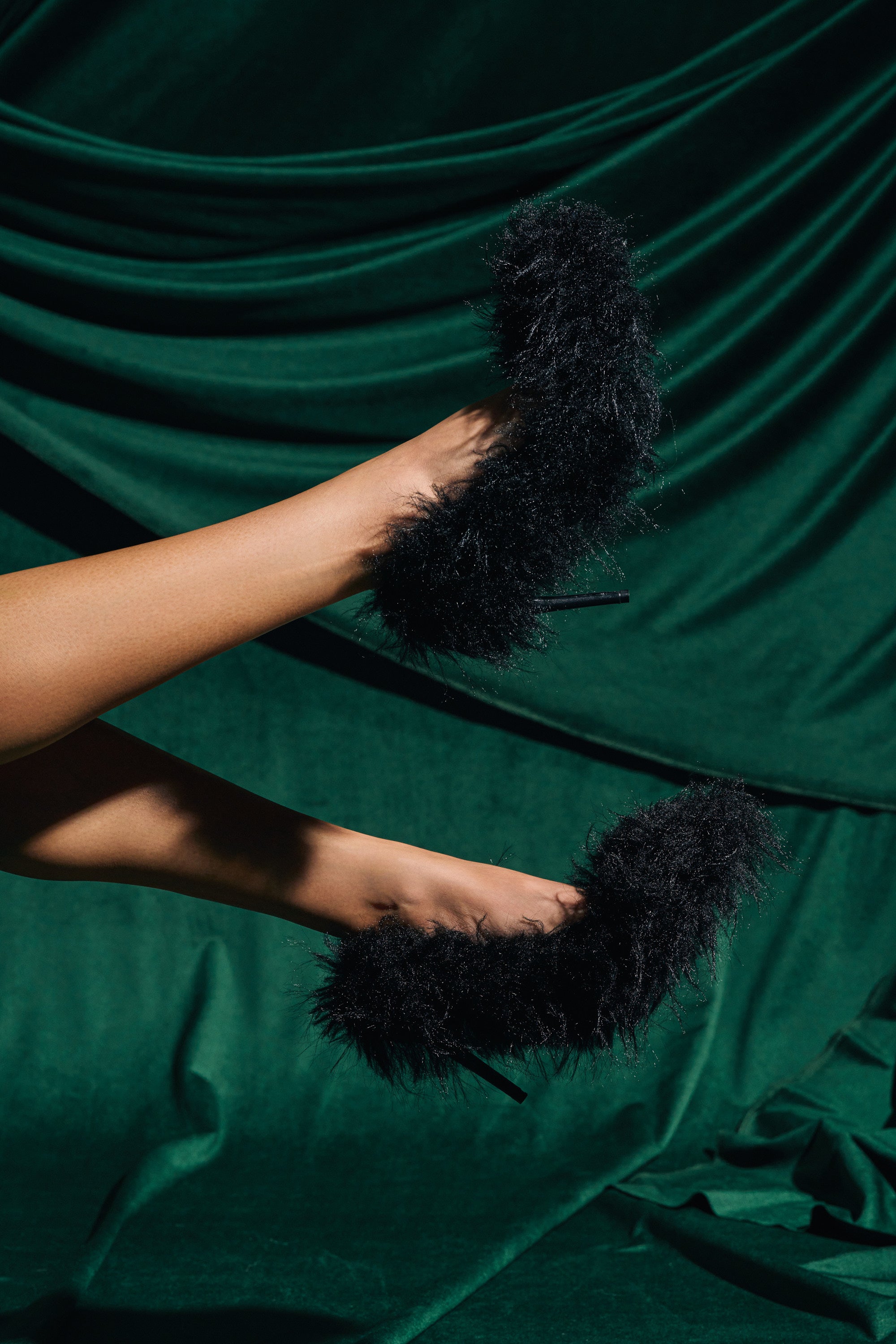 A person's legs are raised, showcasing the AZALEA WANG KURT BLACK FUR EMBELLISHED PUMP with fluffy, textured black faux fur heels against a backdrop of dark green velvet fabric.