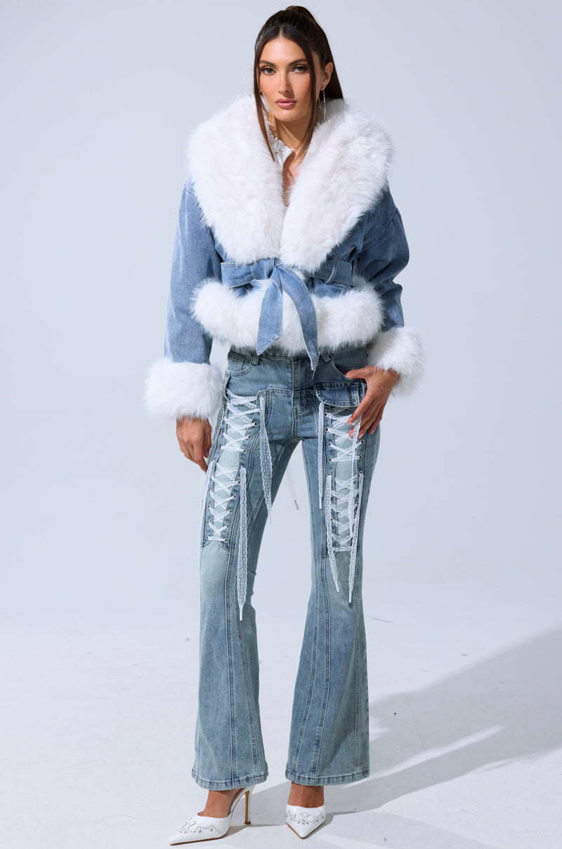 A woman faces forward in the LAINEY FUR TRIM DENIM MOTO jacket with a belt, styled with flared jeans featuring white lace-up details and white pointed-toe heels against a plain light background.