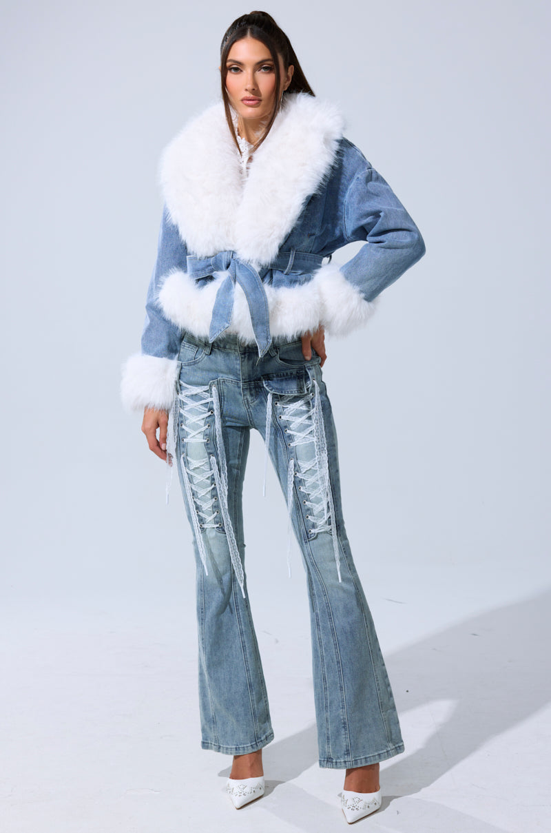 A woman wears the LAINEY FUR TRIM DENIM MOTO, a blue denim jacket with fur trim and a matching tie-waist, paired with flared blue jeans with white lace-up details and white pointed heels. The background is plain light gray.