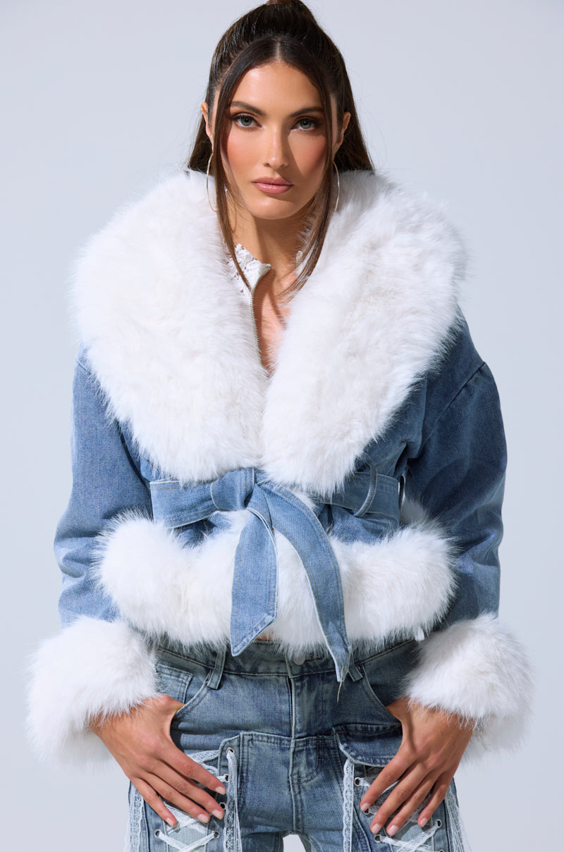 A woman wears the LAINEY FUR TRIM DENIM MOTO jacket with large white faux fur on the collar, cuffs, and hem, belted at the waist. She has long brown hair and stands against a plain light background.