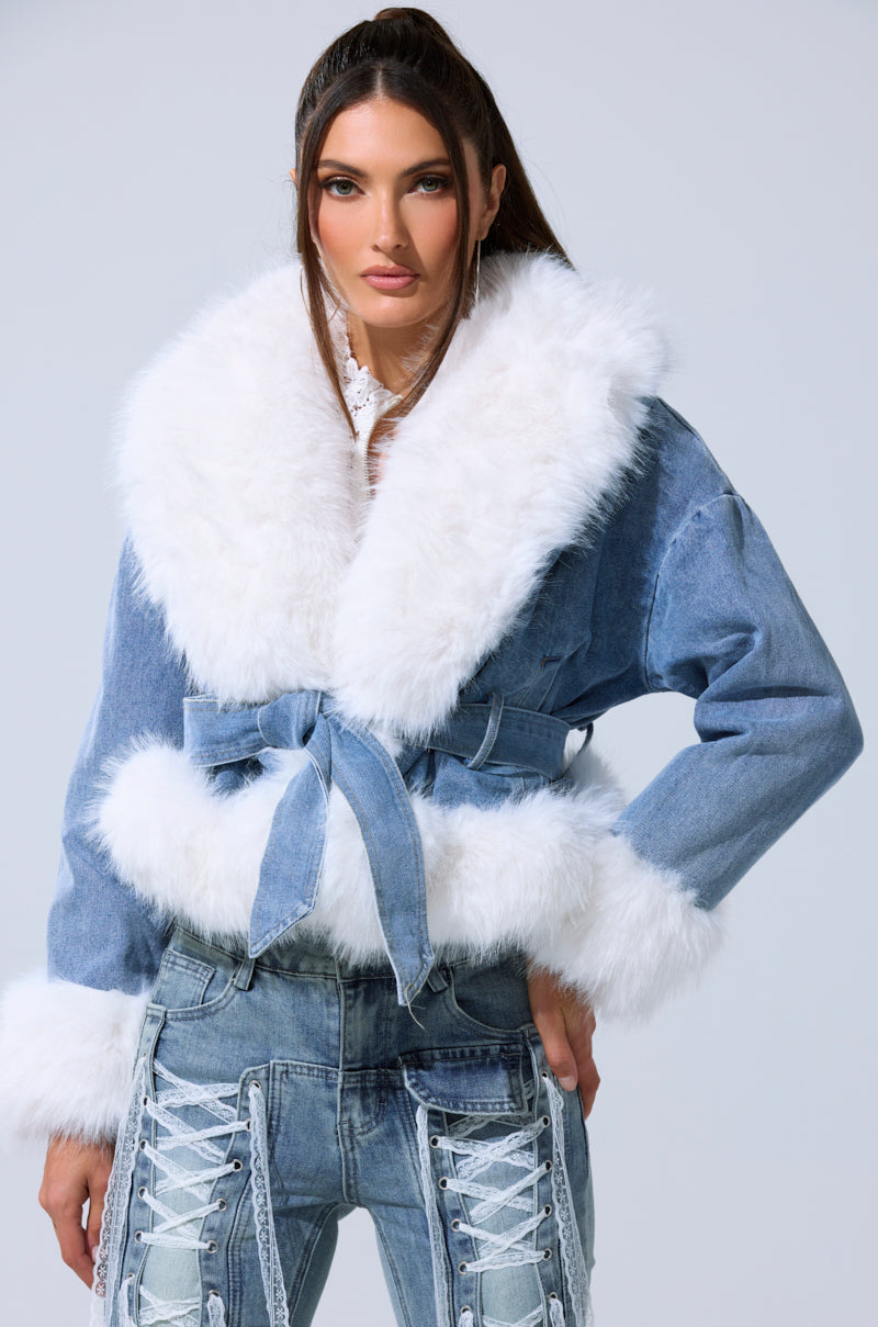 A woman models the LAINEY FUR TRIM DENIM MOTO, a sherpa-lined denim jacket with a wide white faux fur collar, cuffs, and matching belt, styled with light blue lace-up front jeans.