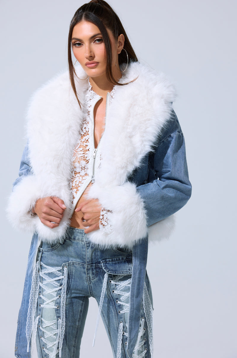 A woman wears the LAINEY FUR TRIM DENIM MOTO—a light blue cropped denim jacket with faux fur trim—over a white lace top and high-waisted, distressed lace-up jeans, standing against a plain light background.