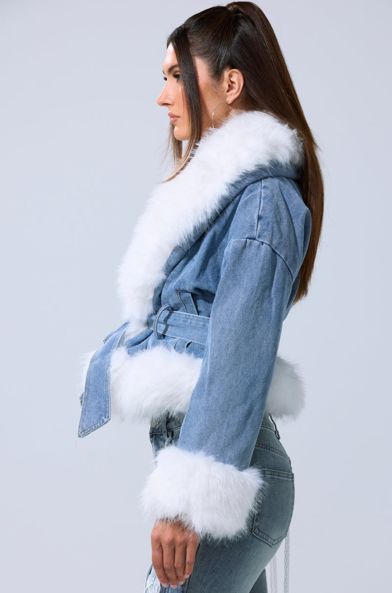 A woman with long brown hair models the LAINEY FUR TRIM DENIM MOTO, featuring white fur trim on the collar, cuffs, and hem. She stands in profile against a plain light background.