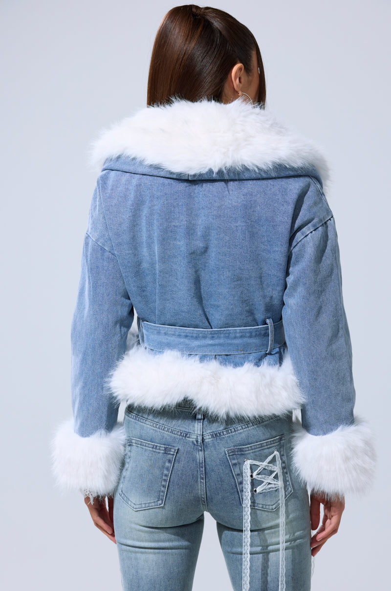 A woman seen from behind wears the LAINEY FUR TRIM DENIM MOTO, a cropped denim jacket with white faux fur trim on the collar, cuffs, and hem, paired with fitted blue jeans featuring lace-up detail on one pocket.