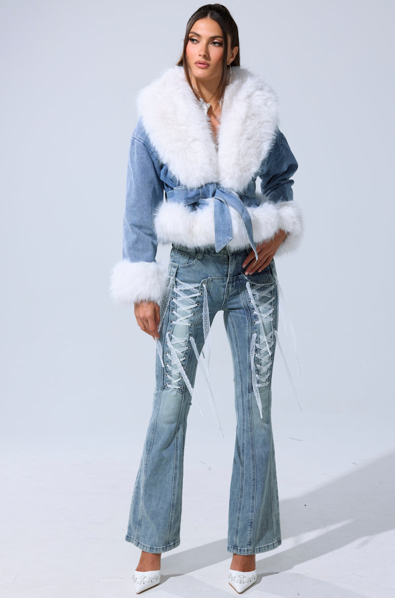 A woman poses against a plain light background wearing the LAINEY FUR TRIM DENIM MOTO, a cropped denim jacket with white faux fur trim and belt, styled with flared lace-up jeans and white heels.