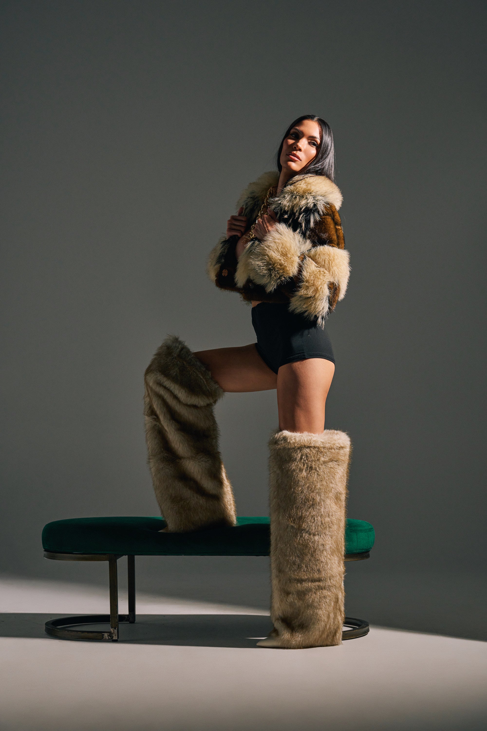 Wearing the AZALEA WANG LAGANN BONE THIGH HIGH FUR LUXE BOOT, a woman poses confidently with one foot on a green bench, dressed in a fur jacket and black bodysuit under dramatic lighting against a plain backdrop.