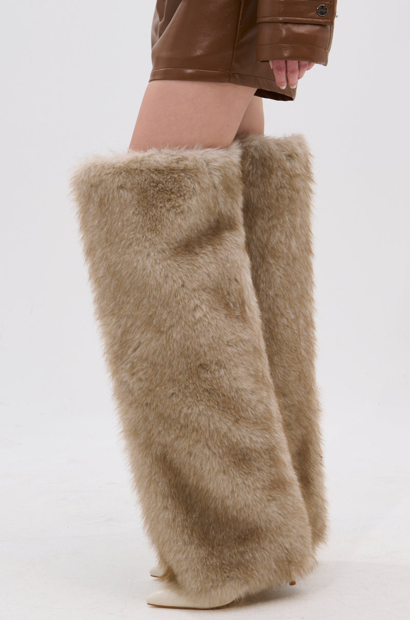A person wears AZALEA WANG LAGANN BONE THIGH HIGH FUR LUXE BOOTS over stiletto heels, styled with a brown leather skirt and jacket. The chic outfit stands out against a plain white background.