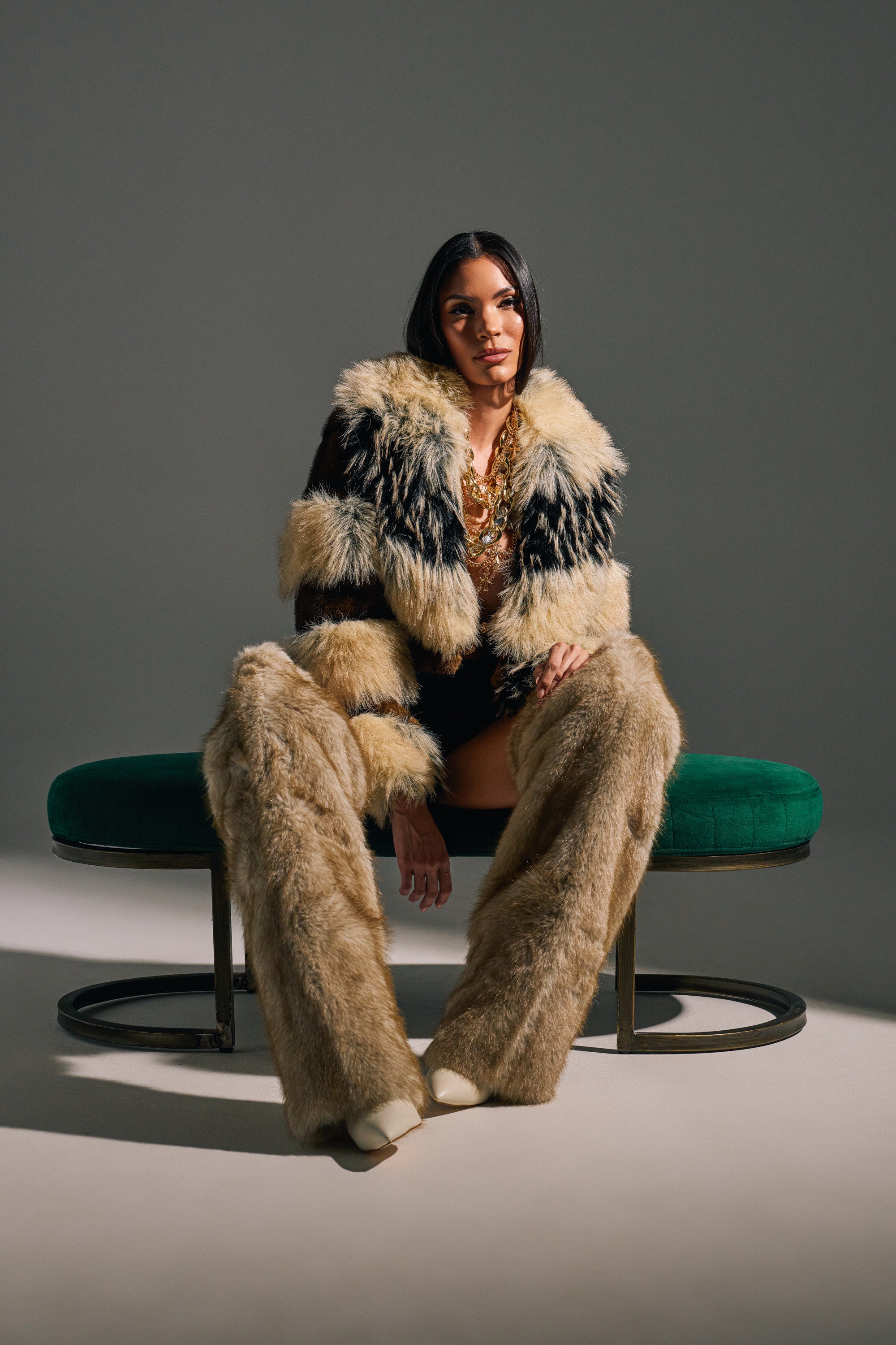 A woman with long dark hair sits on a green bench in a black top, tan and black faux fur coat and pants, and AZALEA WANG LAGANN BONE THIGH HIGH FUR LUXE BOOTS. She wears layered necklaces, has a neutral expression, and soft lighting surrounds her.