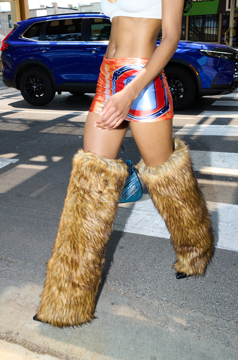 Someone walks down a city street in the AZALEA WANG LANGDON BROWN THIGH HIGH FUR LUXE BOOT, paired with orange and blue shorts and a white crop top, with a blue SUV visible in the background.