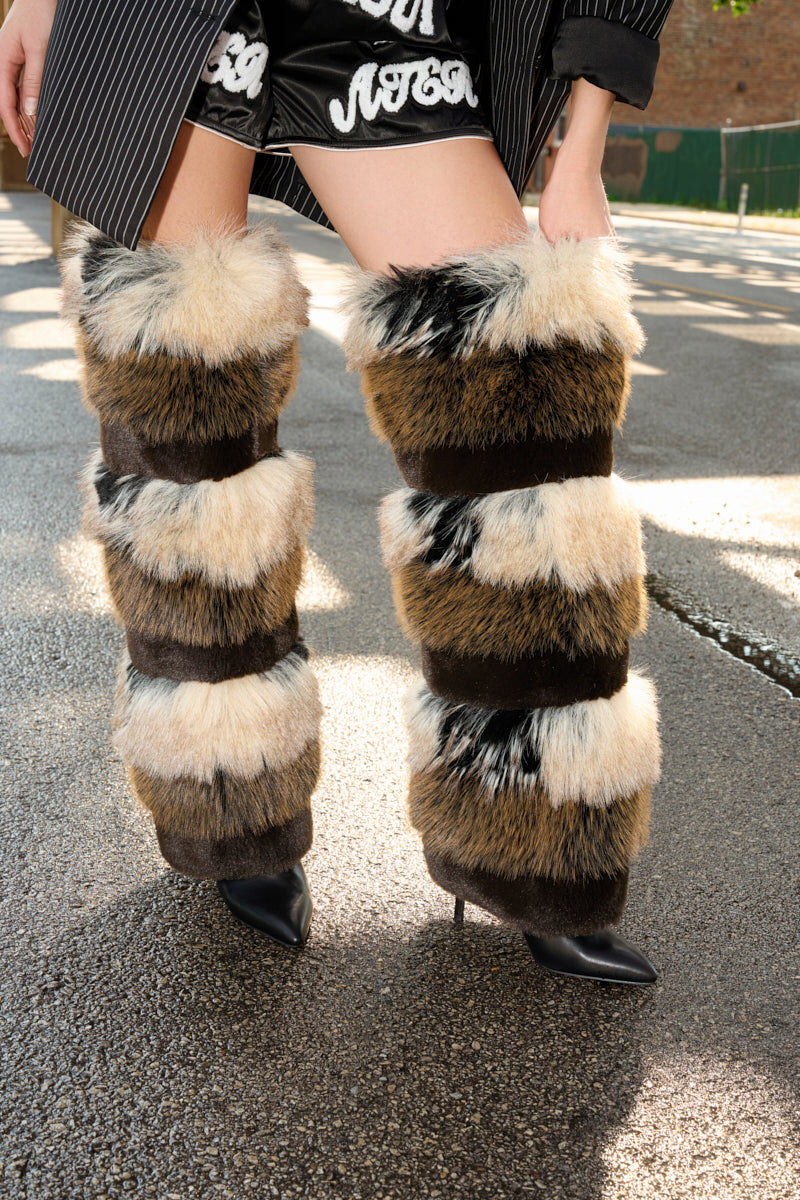 A person wearing black shorts, a striped coat, and AZALEA WANG LANGLAU BROWN MULTI STILETTO FUR BOOT with cream, brown, and black stripes stands on wet pavement, the striking boots elevating the bold outfit.