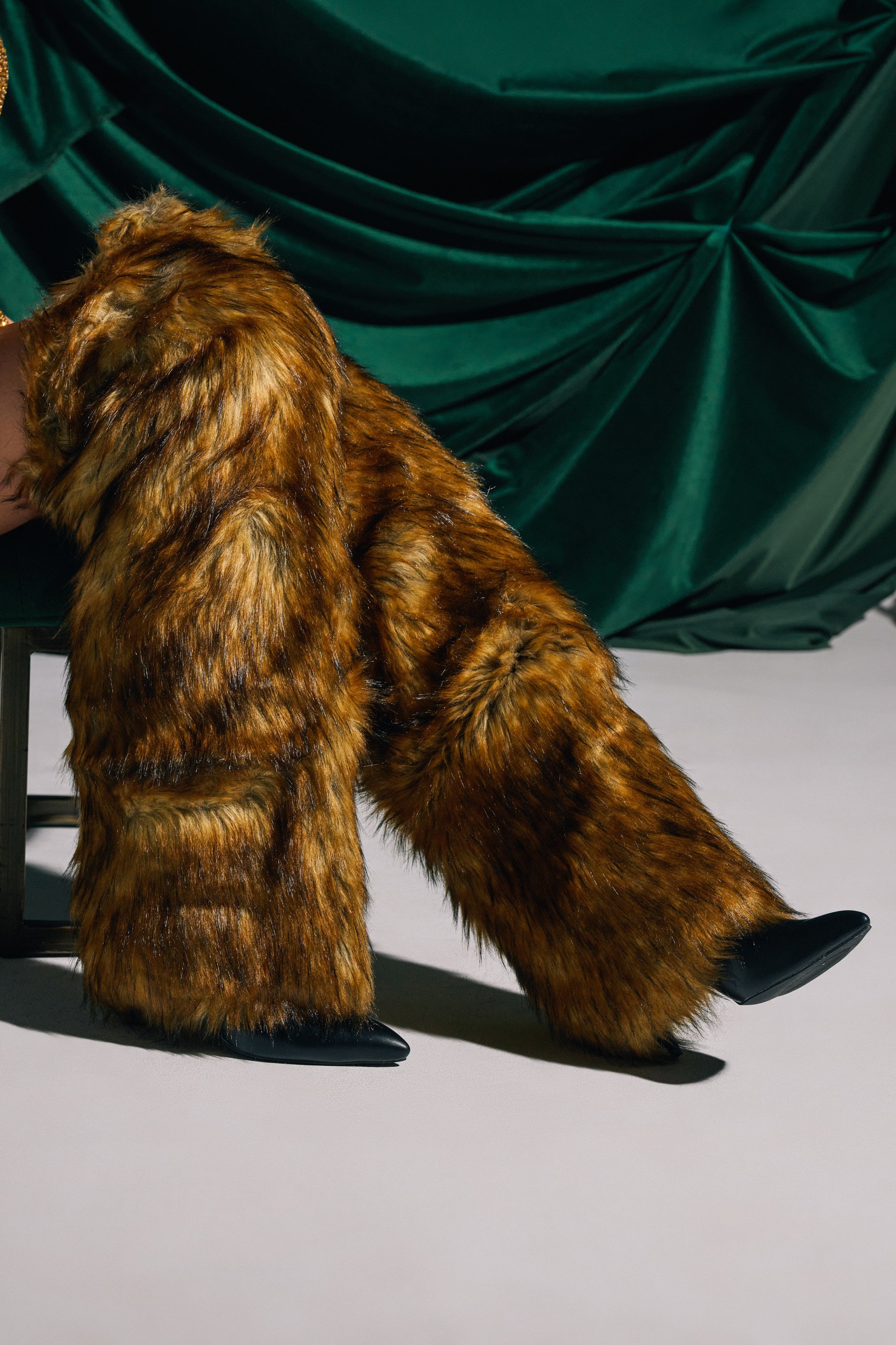 A person seated against a green draped fabric background wears the AZALEA WANG LANGLEYE BROWN THIGH HIGH FUR LUXE BOOT with brown faux fur pants and sleek stiletto heels.