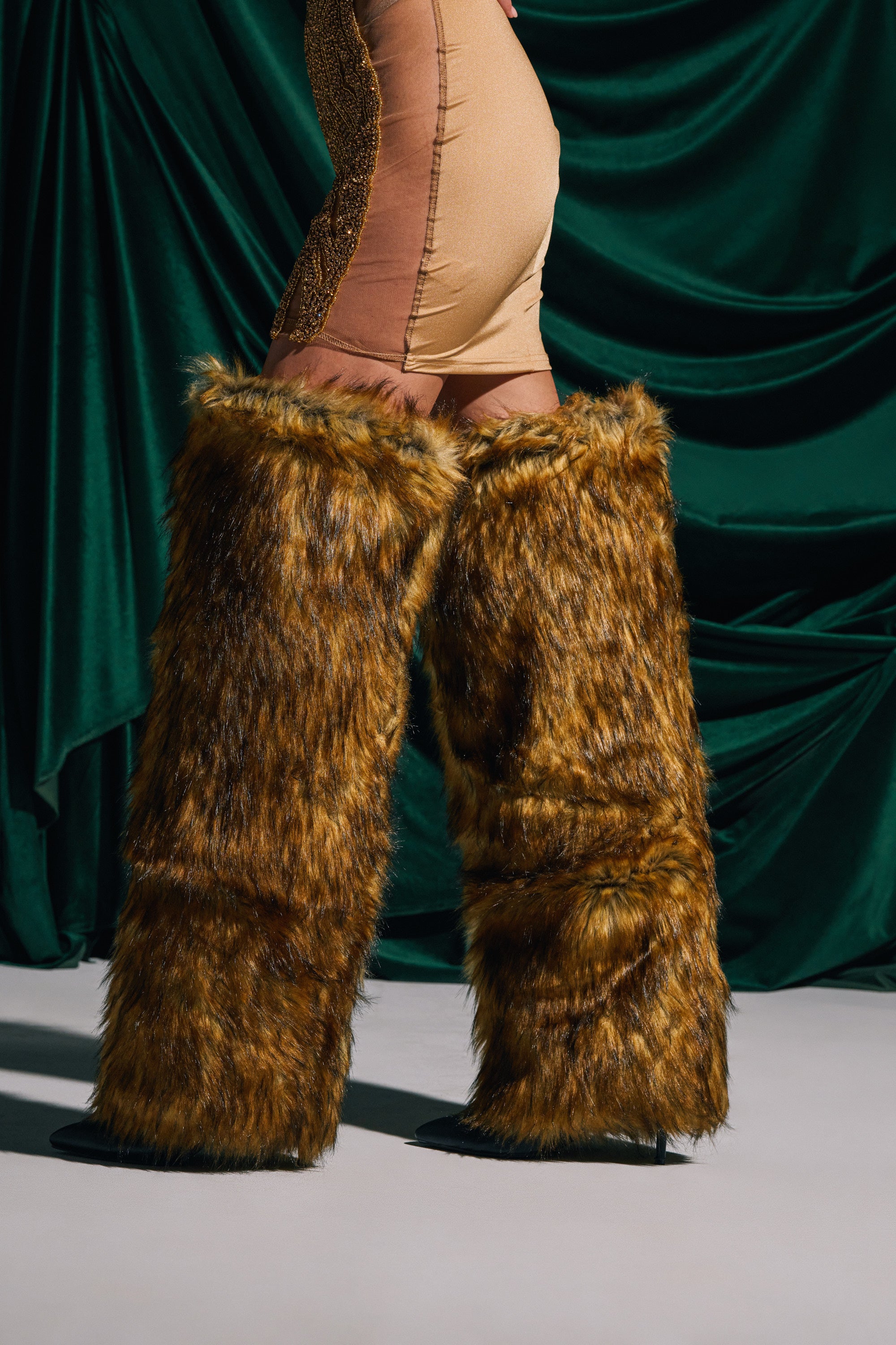 Wearing AZALEA WANG LANGLEYE BROWN THIGH HIGH FUR LUXE BOOT with a stiletto heel and tan leggings with gold details, person stands before dark green draped fabric—only their legs visible.