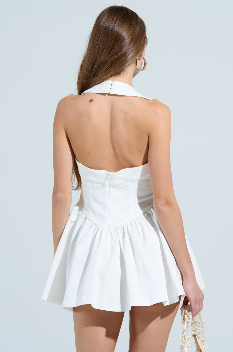 A person with long brown hair, seen from the back, wears a Rosalia Stretch Linen Mini Dress in white featuring sleeveless design and halter straps. The fitted bodice complements the flared skirt as they hold a small textured bag against a plain light gray background.