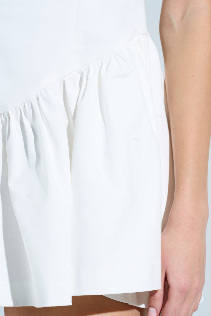 Close-up shot of someone in the ROSALIA STRETCH LINEN MINI DRESS IN WHITE, emphasizing its fitted bodice and gathered skirt. The fabric's texture and draping are highlighted, with part of their arm visible.