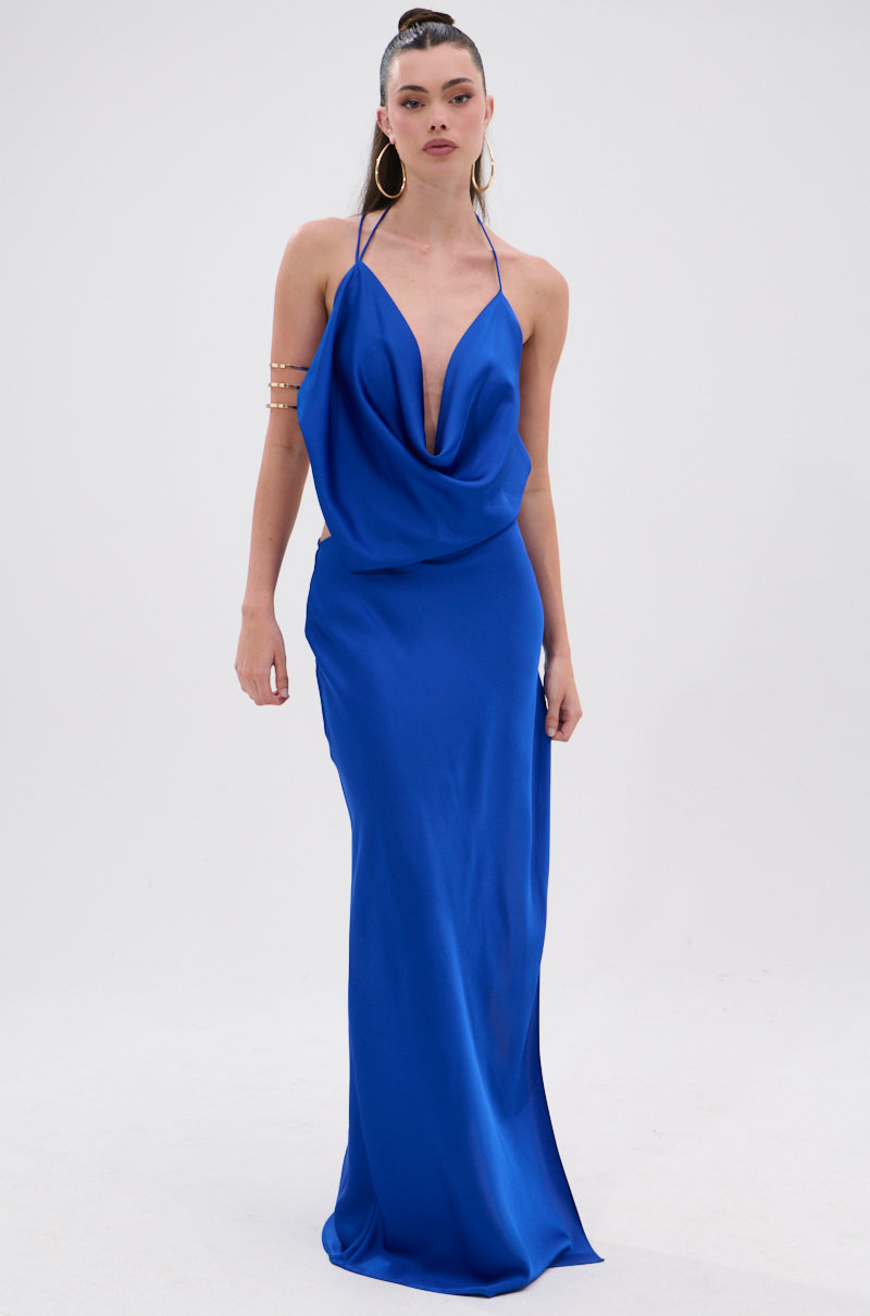 A woman stands against a white background wearing the RAYNA SATIN MAXI DRESS IN COBALT BLUE with a plunging cowl neckline, hoop earrings, and a gold arm cuff. Her hair is styled in a high ponytail.