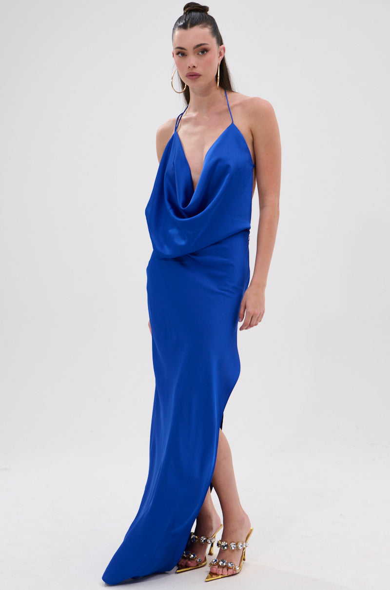 A woman wears the RAYNA SATIN MAXI DRESS IN COBALT BLUE featuring a plunging cowl neckline, halter neck, and high side slit, styled with jeweled heels and gold hoop earrings against a plain white background.