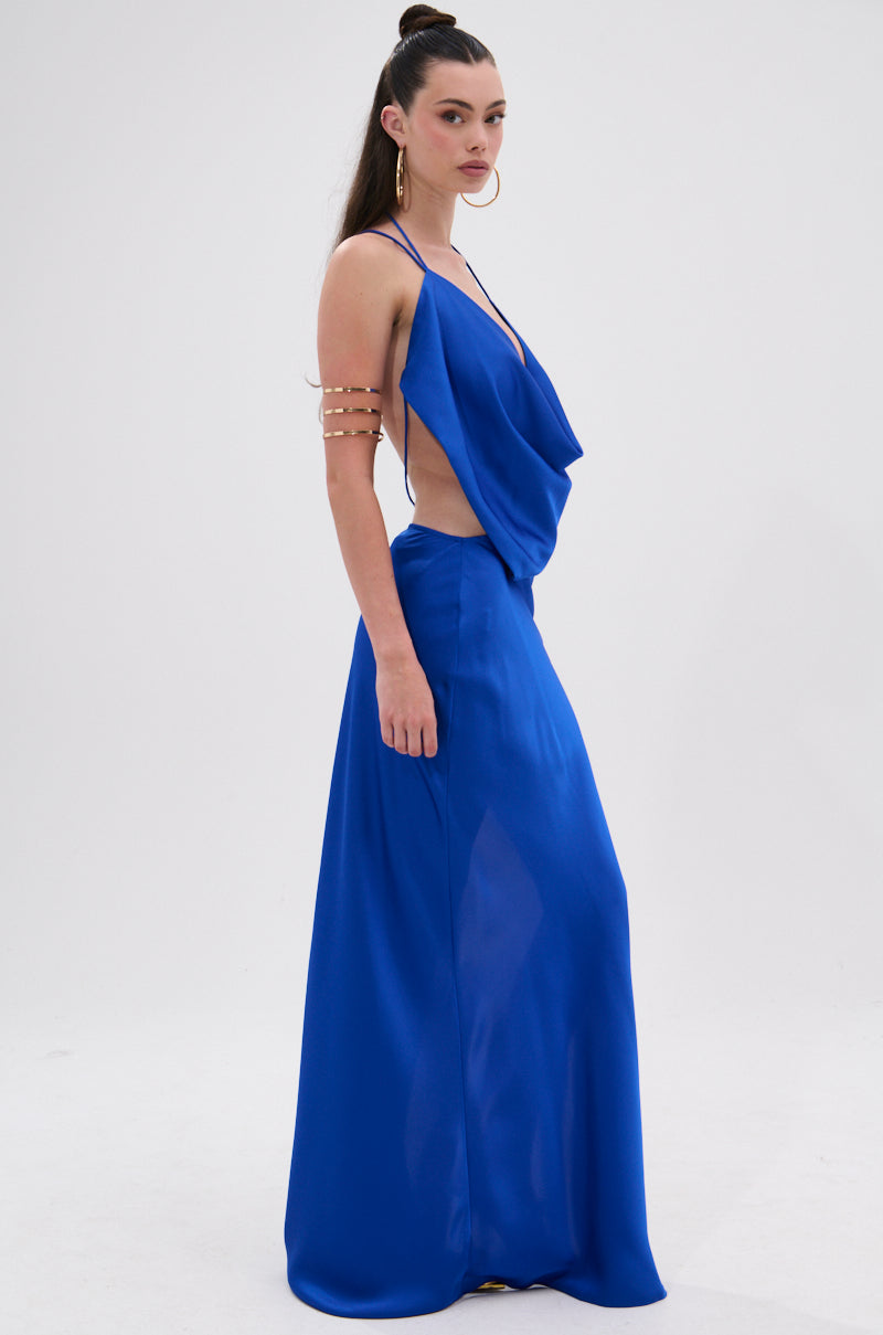 A woman in profile wears the RAYNA SATIN MAXI DRESS IN COBALT BLUE, featuring a plunging cowl neckline and open sides. She styles her long dark hair in a high ponytail and accessorizes with a gold arm cuff against a plain white background.