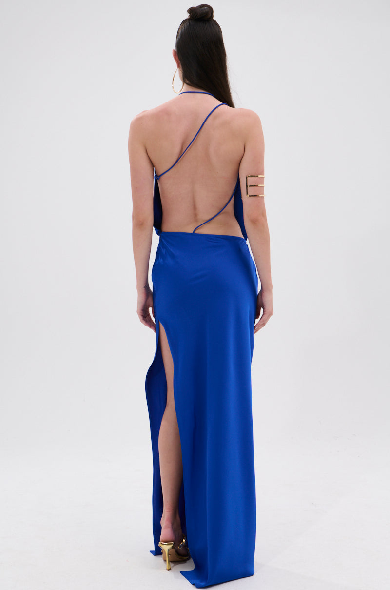 A woman faces away, wearing the RAYNA SATIN MAXI DRESS IN COBALT BLUE featuring thin straps, a plunging cowl neckline, and a high slit. Her long dark hair is in a top knot, paired with hoop earrings and gold heels.
