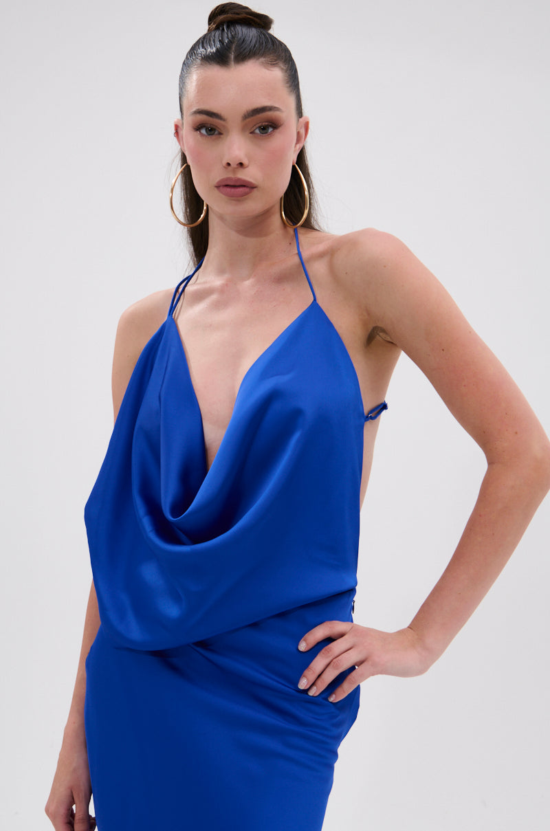 A woman wears the RAYNA SATIN MAXI DRESS IN COBALT BLUE, featuring a sleeveless design, spaghetti straps, and a plunging cowl neckline. She stands against a plain light background with hoop earrings, hair pulled back, and one hand on her hip.