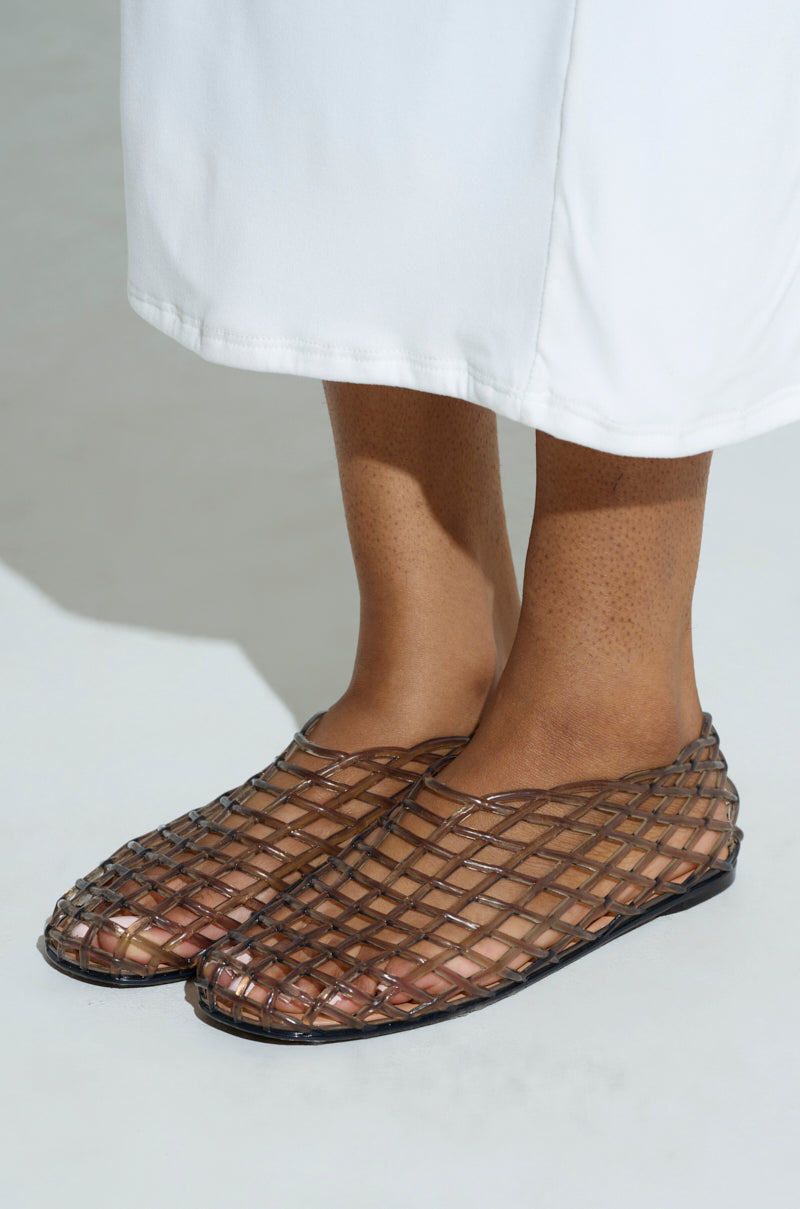 A person stands on a light surface, wearing sheer woven AKIRA LINUS BLACK FLAT slip-ons with an almond toe and net-like pattern. Their legs are covered by a calf-length white garment.