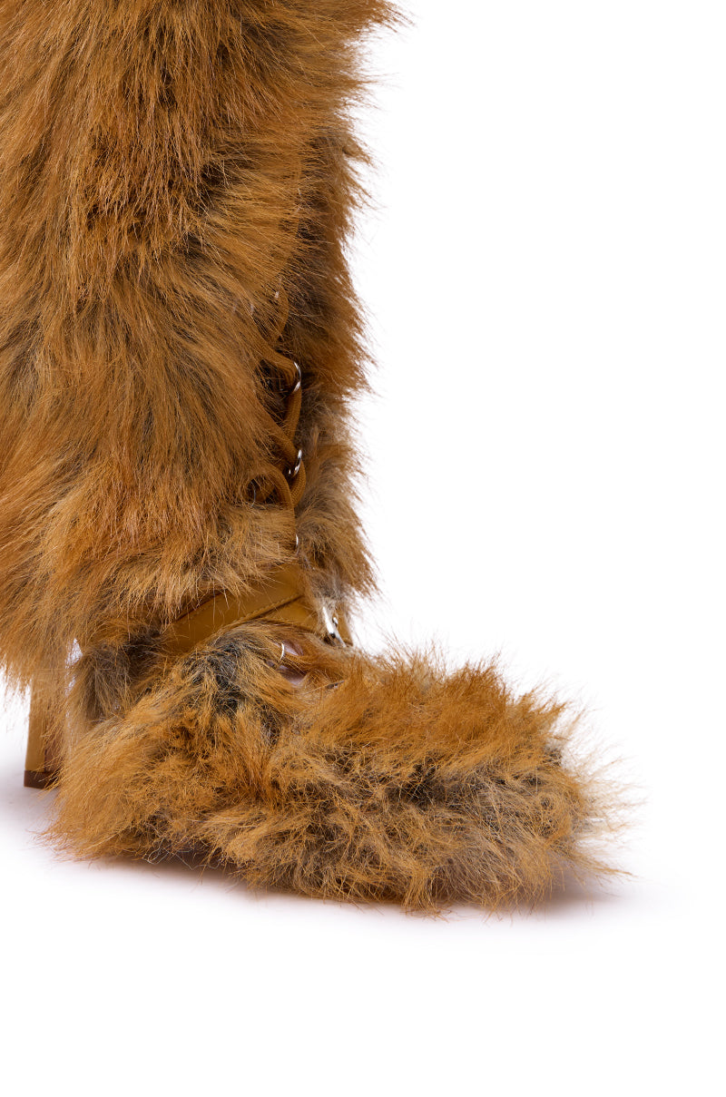 A close-up of a person wearing the AZALEA WANG LOURDANNE TAN FUR LACE UP STILETTO BOOT, featuring shaggy brown faux fur and partially visible laces, against a plain white background.