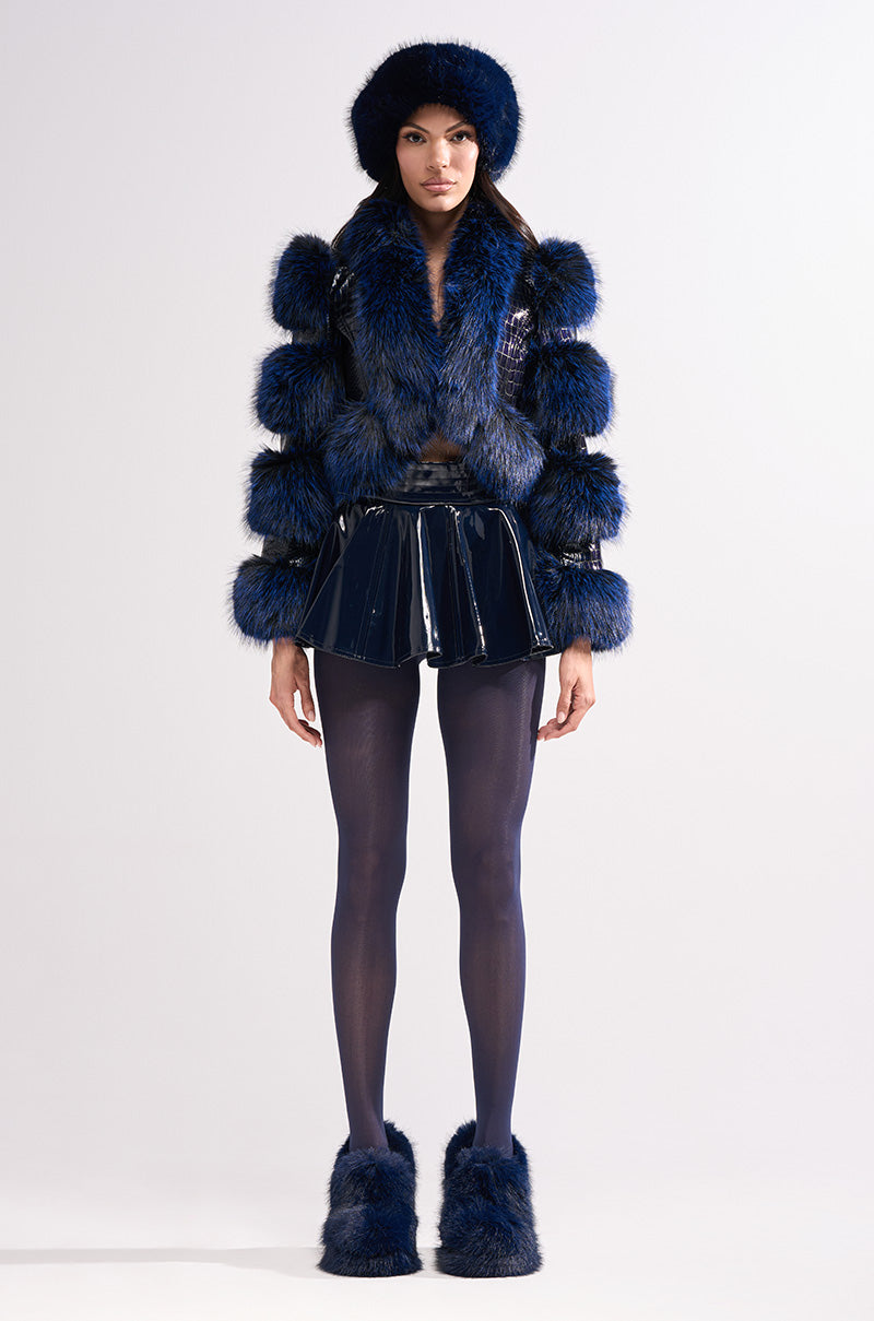 A woman faces forward in the LOVERBOY FUR TRIM MOTO IN NAVY CROC jacket with fur trim, paired with a shiny mini skirt, dark tights, and blue fur slippers against a plain light background.