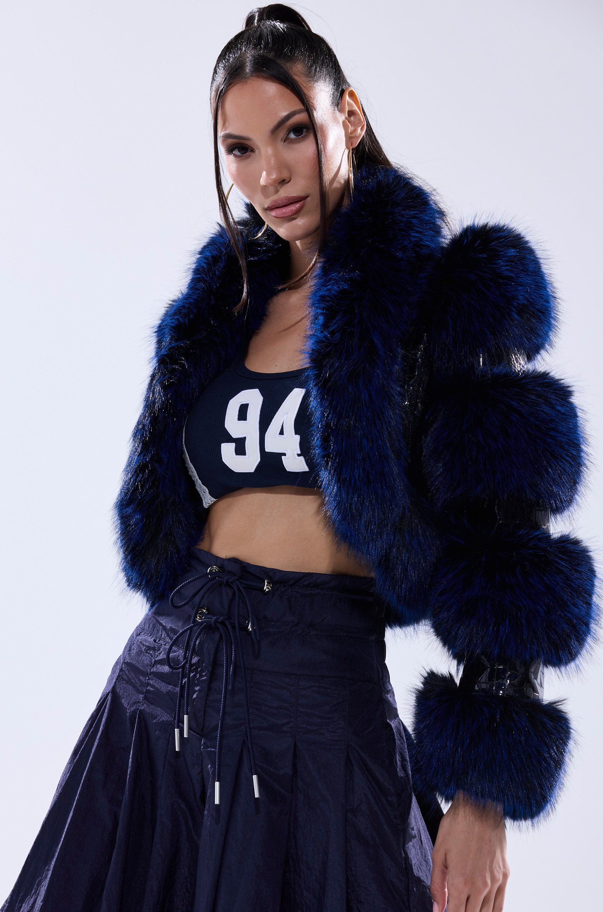 A woman stands confidently against a white background wearing the LOVERBOY FUR TRIM MOTO IN NAVY CROC, a navy sports bra with "94" printed on it, and a dark pleated skirt.