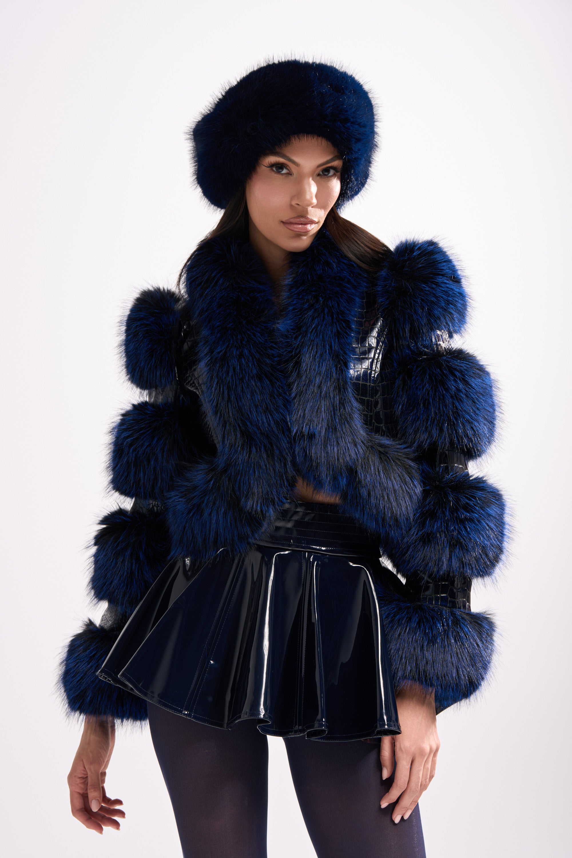 A woman models the LOVERBOY FUR TRIM MOTO IN NAVY CROC, featuring fluffy panels and paired with a matching faux fur hat, shiny dark blue skirt, and black tights, all set against a plain light background.