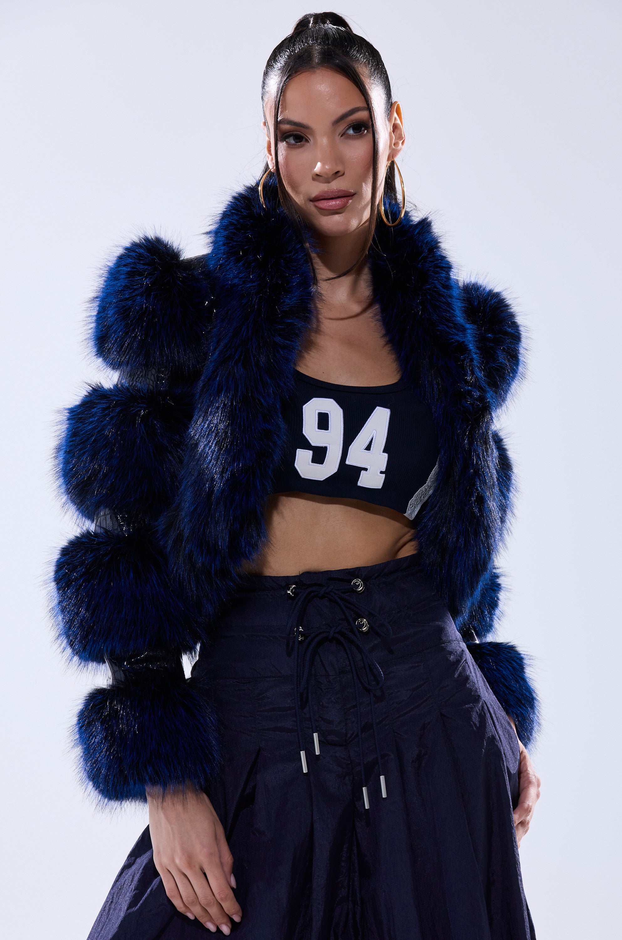 A woman stands confidently against a plain, light background, wearing high-waisted navy pants, a navy crop top with the number 94, and the LOVERBOY FUR TRIM MOTO IN NAVY CROC jacket.