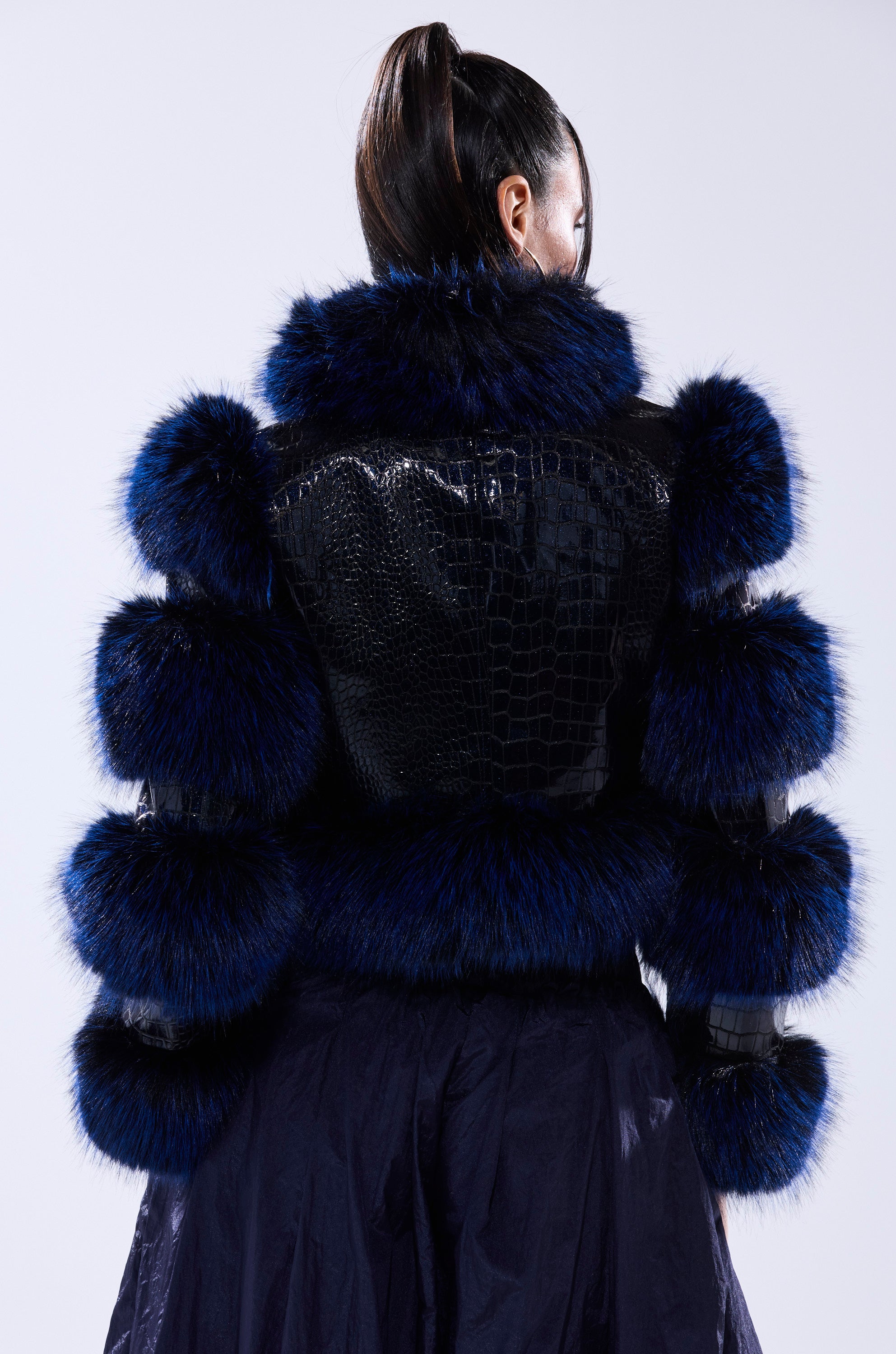 A person with long hair in a ponytail wears the LOVERBOY FUR TRIM MOTO IN NAVY CROC, paired with a dark skirt, shown from the back against a light background.