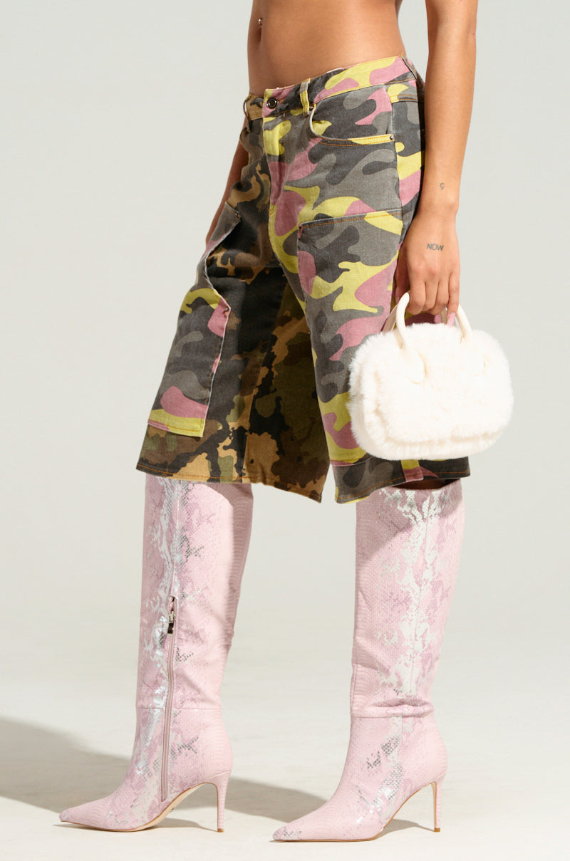 A person wearing GO TIME CAMO SHORT utility-style camouflage knee-length cargo shorts, pink knee-high boots with zippers, and holding a small fluffy white purse. Part of their midriff and tattooed arm are visible against a plain background.
