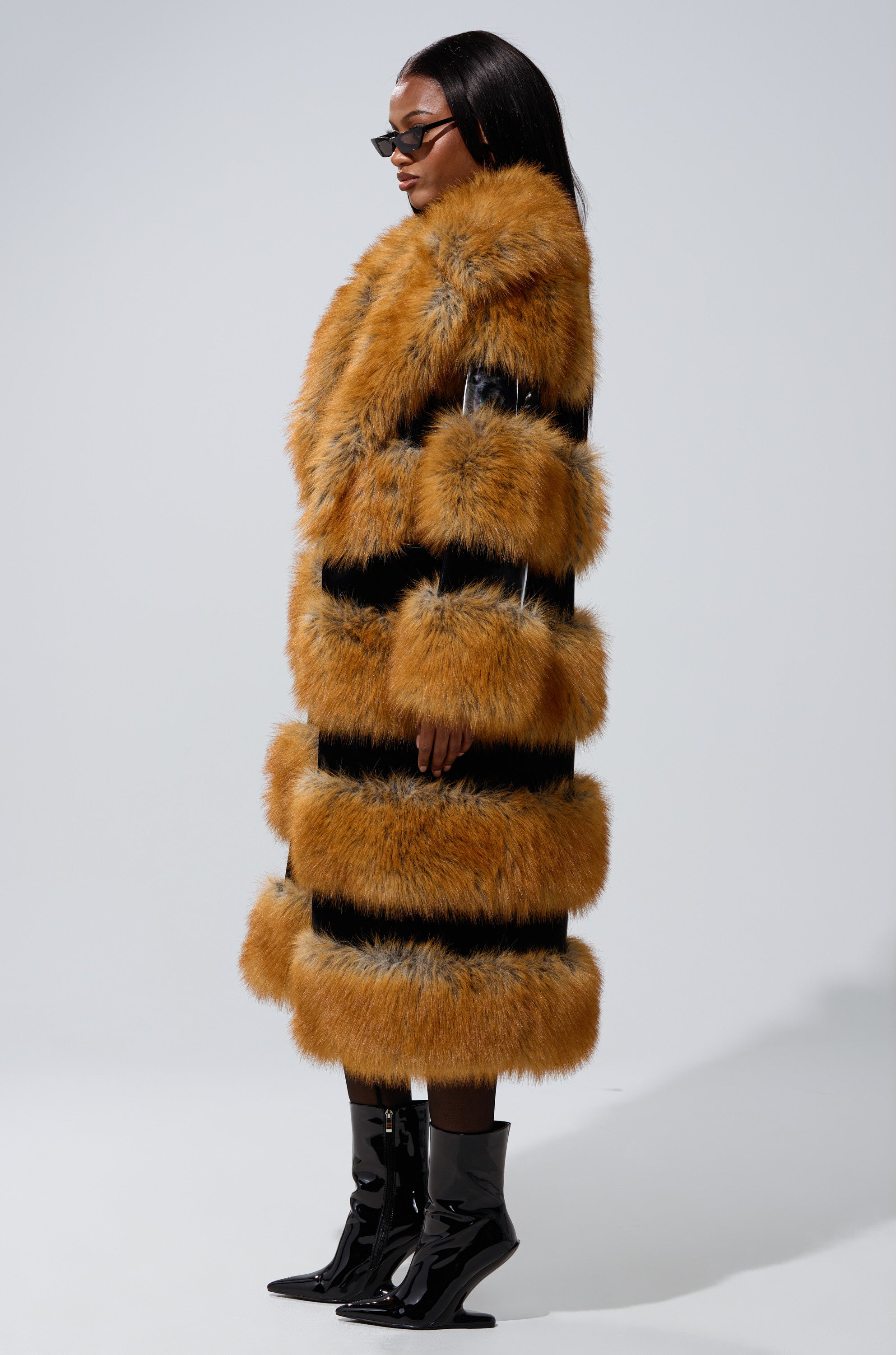 Wearing the MADDOX PANELED FAUX FUR coat with bold orange-brown and black panels, sunglasses, and black boots, a person stands confidently against a plain grey background.