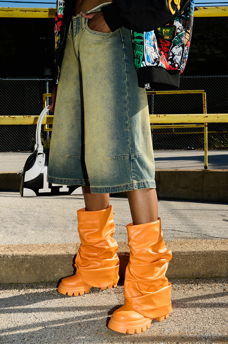 A person dressed in wide-legged denim shorts, orange AZALEA WANG MATE booties featuring a chunky heel, and a colorful graphic jacket stands on the sidewalk beside a black and white bag, with a yellow railing and chain-link fence visible in the background.