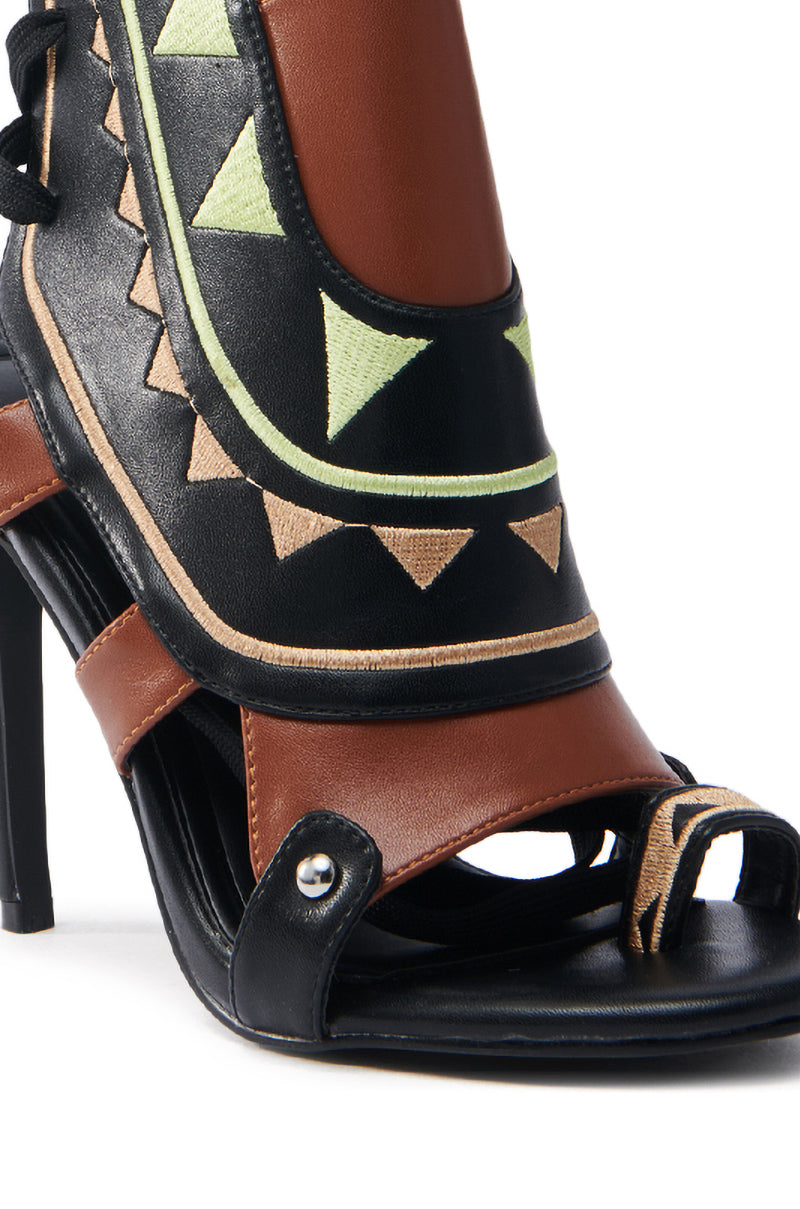 Close-up of the AZALEA WANG MERAKI BLACK AND BONE SANDAL featuring a color block design with a black base, brown accents, and geometric patterns in green and tan triangles on the vegan leather upper. The sandal has a strap embellished with metal studs.