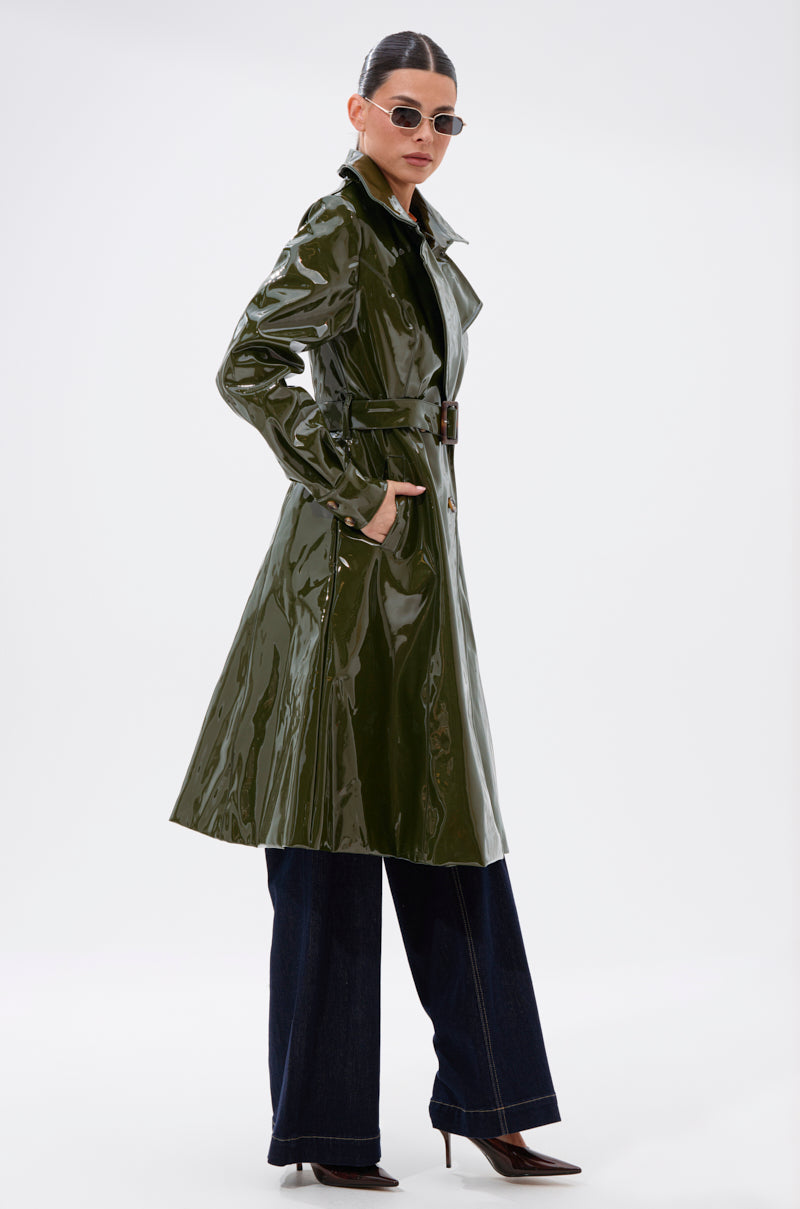 A woman wearing the MITICA PATENT TRENCH IN OLIVE, dark wide-leg jeans, brown heeled boots, and sunglasses stands confidently against a plain white background.