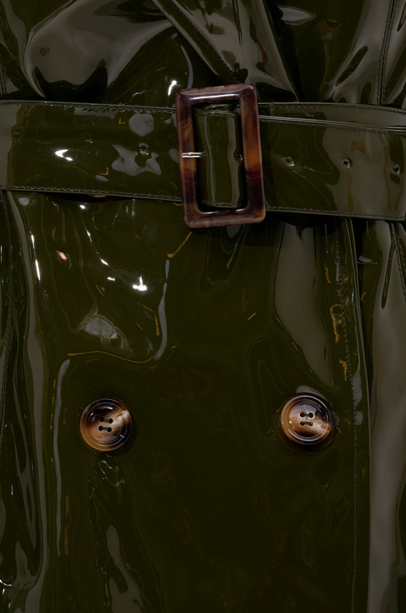 Close-up of the MITICA PATENT TRENCH IN OLIVE, a shiny dark green faux patent leather trench coat with a wide belt, rectangular tortoiseshell buckle, and two large brown marbled buttons.